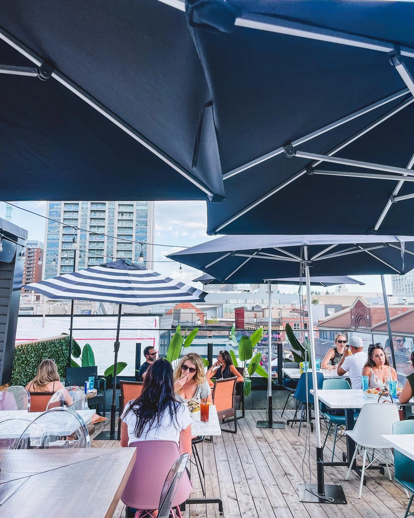 People dining at an outdoor rooftop restaurant with umbrellas, surrounded by city buildings. rooftop, full bar, full menu, dinner, views, sunsets. best rooftop in Ottawa