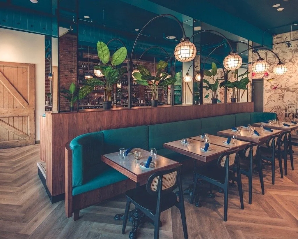 A cozy restaurant interior with wooden tables, green upholstered seating, potted plants, and decorative hanging lights. a timeless building in Ottawa, one of the best events venues, great pricing, amazing food, beautiful decor, outstanding service