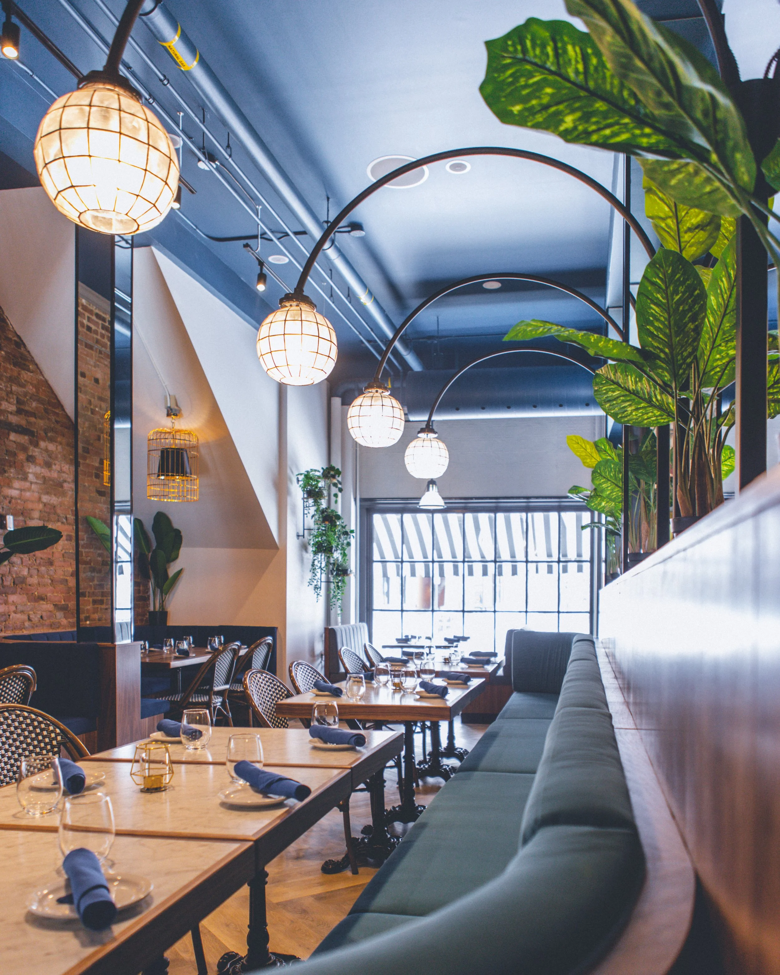 Interior of a modern restaurant with wooden tables set with wine glasses, black napkins, and small candle holders. There are black and white rattan chairs and a long cushioned bench along the right side. Hanging globe and cage pendant lights illumina