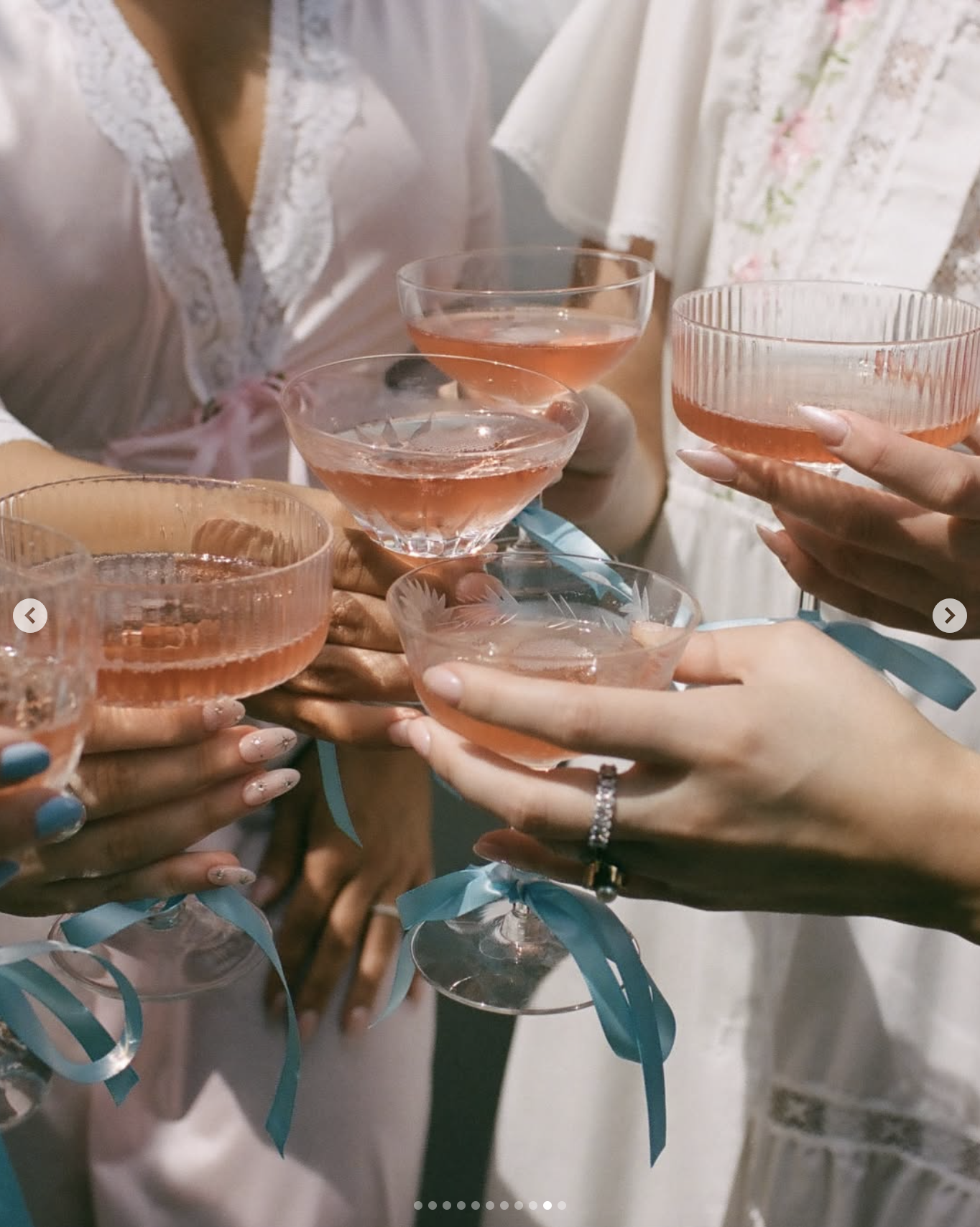 Multiple hands holding champagne glasses with pink beverages, celebrating together, some hands have rings, while others have painted nails. The background shows people in wedding or formal attire.