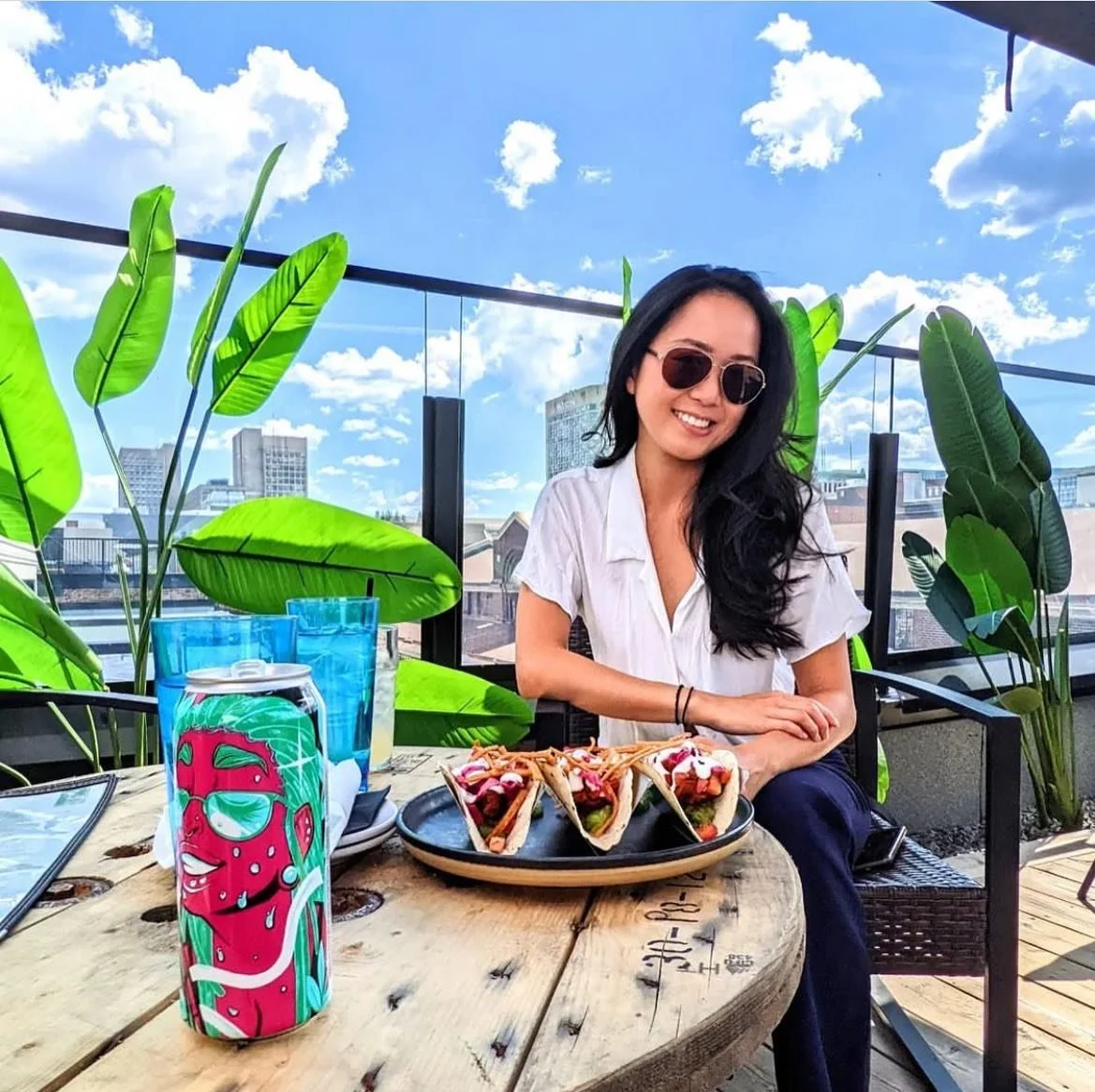 A woman with long dark hair and sunglasses sitting at a rooftop restaurant table with a clear sky, city skyline, and green plants in the background. She is smiling at the camera next to a plate of three tacos and a colorful drink can.