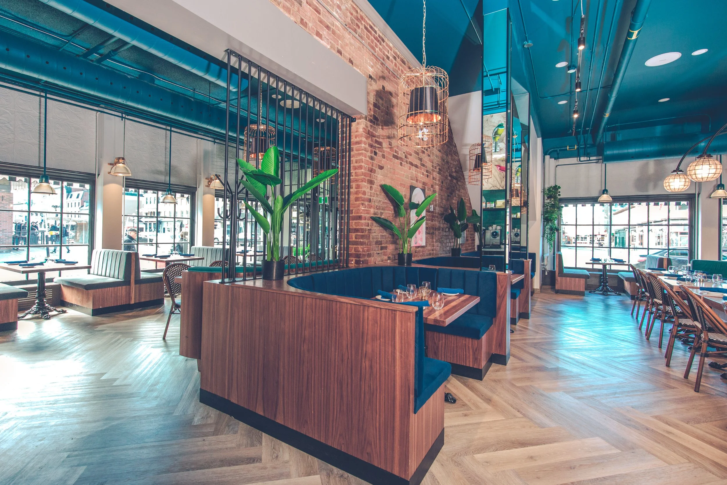 Inside a modern restaurant with exposed brick walls, large windows, and wooden flooring. Mi seating arrangements, including banquettes and chairs, with large potted plants and hanging light fixtures. Heritage building from 1875 | Ottawa Venues