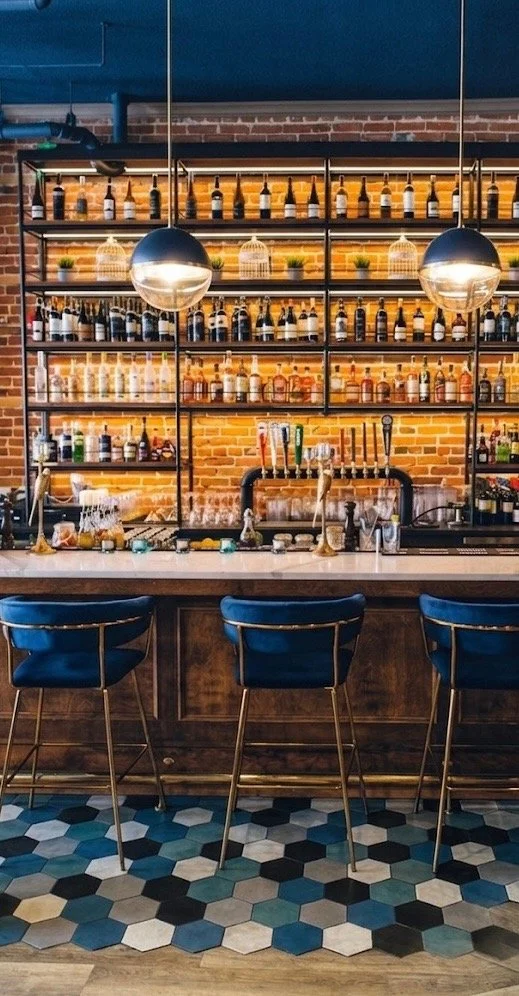 A modern bar with a brick wall behind the shelves filled with bottles, potted plants, and glassware. There are three blue bar stools with gold accents in front of a counter with taps. The floor has a colorful hexagon tile pattern.