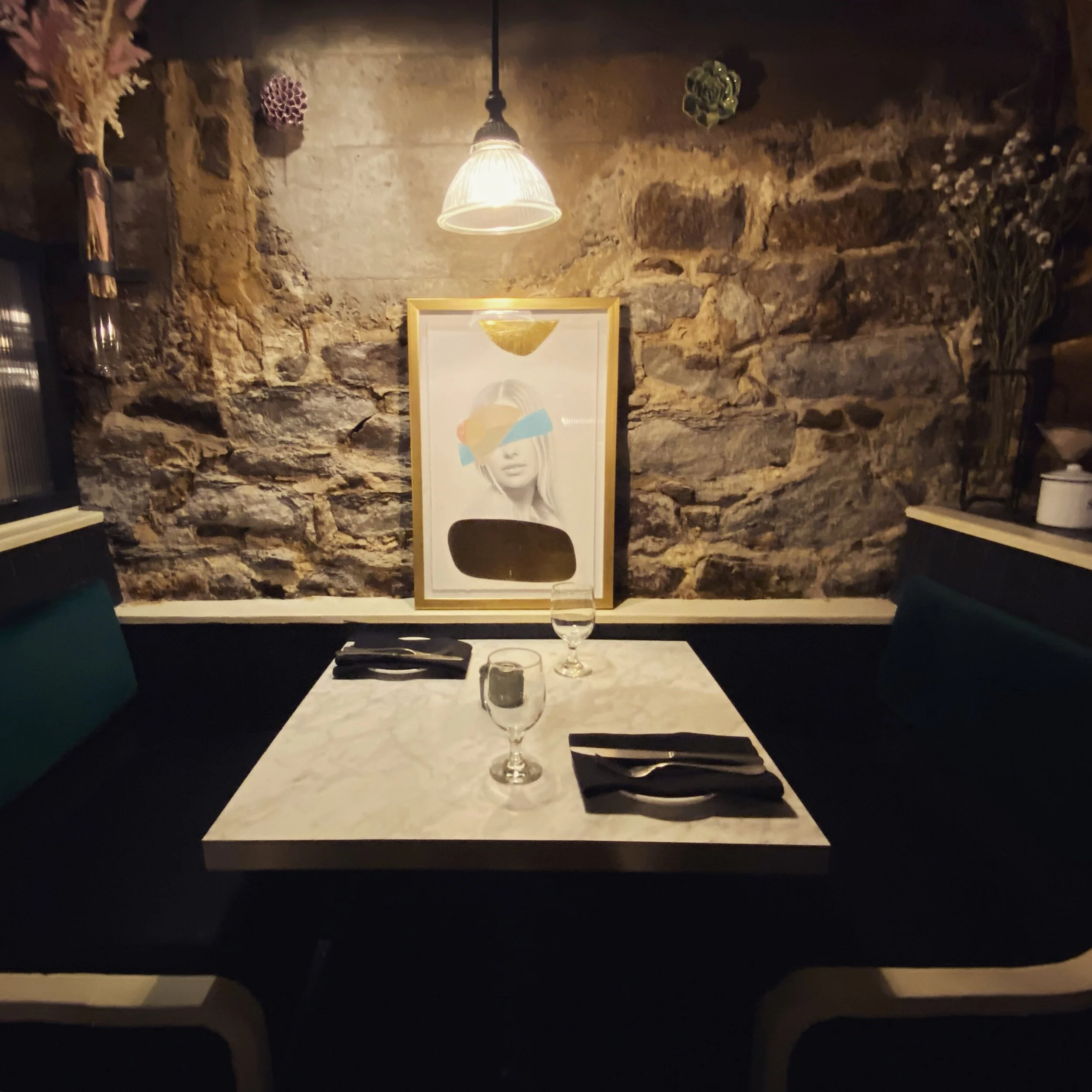 A cozy dining table set for two with black napkins, glasses, and a small candle, against a rustic stone wall with artwork and decorations in the background. Ottawa venues, apothecary, first speakeasy with a story 