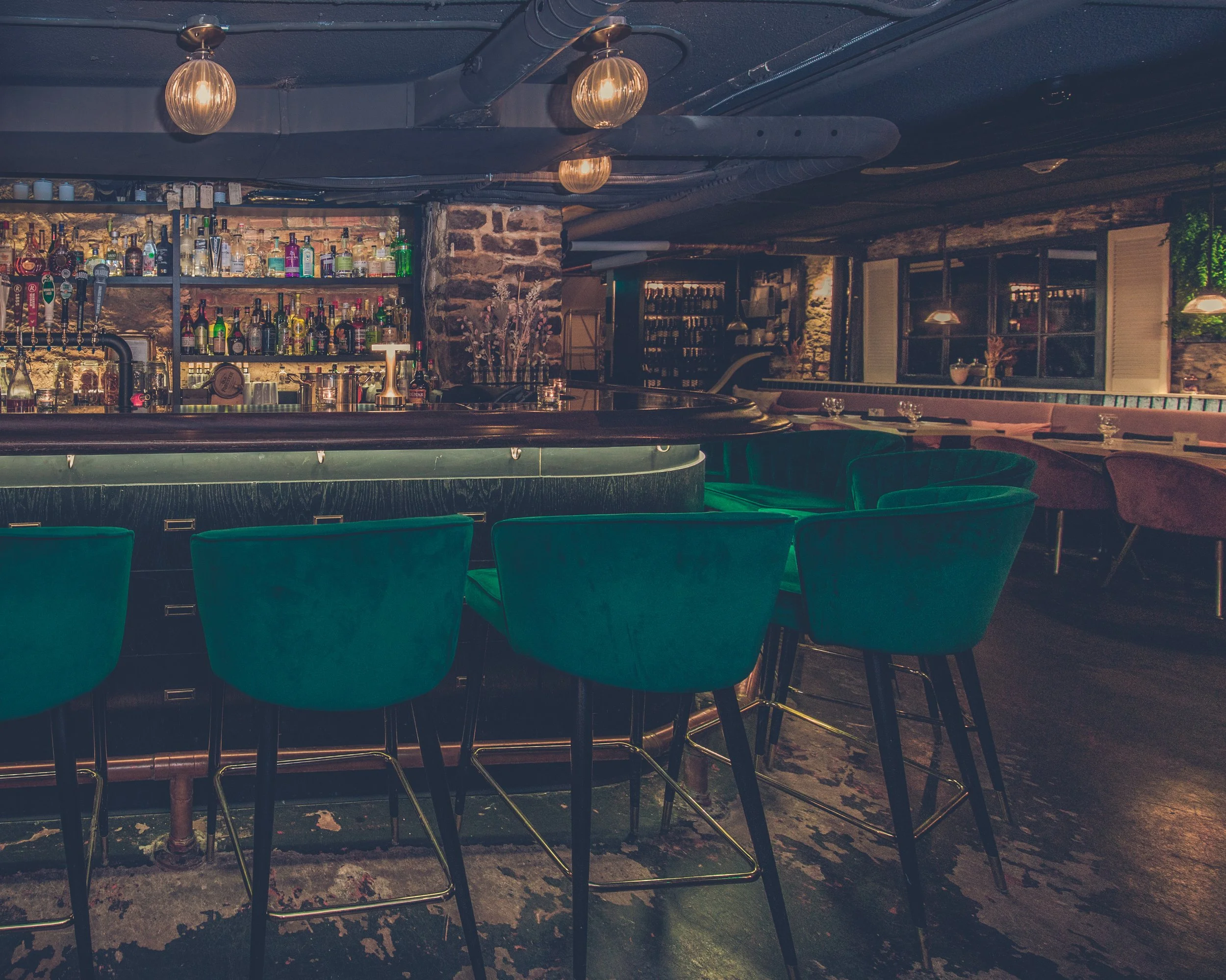 A bar with a stone wall behind it, shelves stocked with various bottles of liquor, a counter with a candle, and teal chairs arranged in front. The ceiling has industrial pipes and hanging lights. heritage building, timeless setting, hand-crafted 