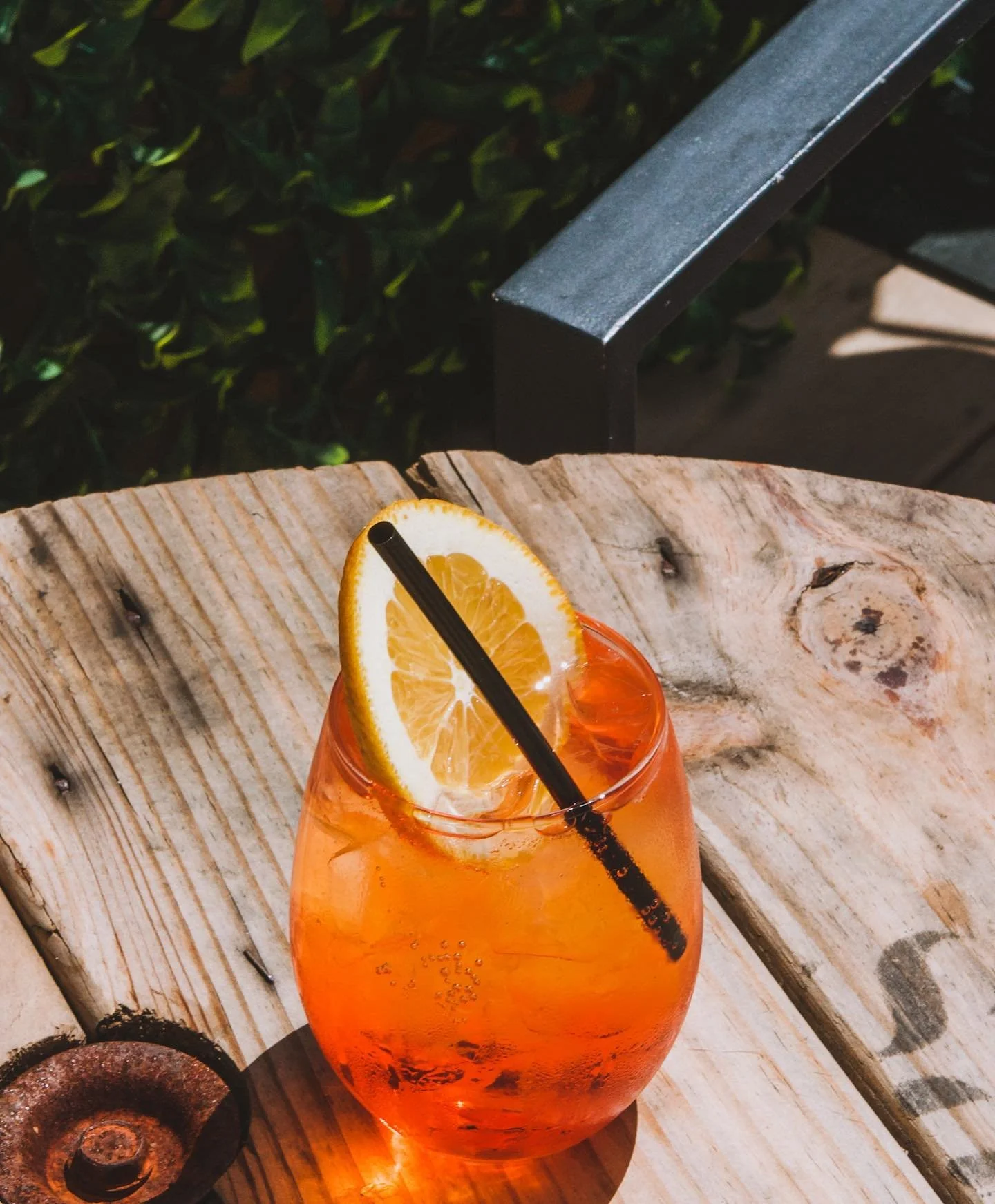 A glass of orange-colored beverage with a lemon slice and a black straw on a wooden surface outdoors.