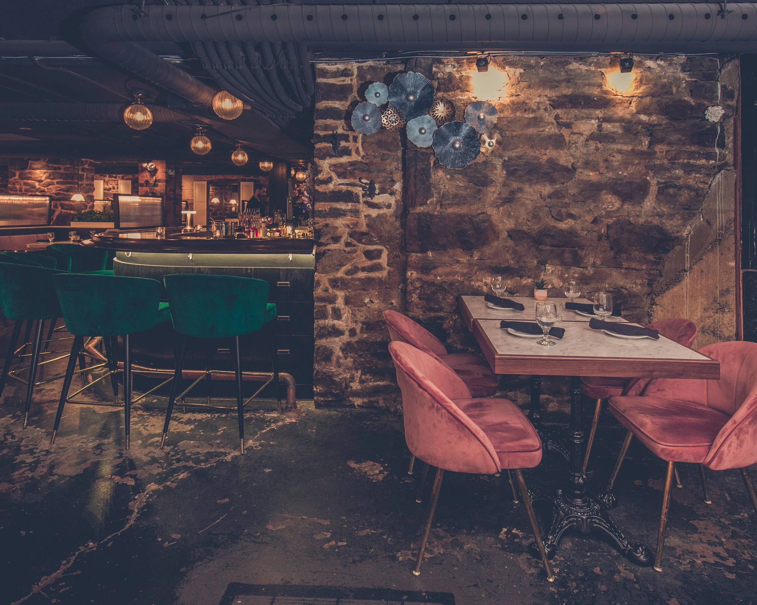 A cozy restaurant with exposed brick walls, velvet pink chairs, a marble-top table set with wine glasses and black napkins, decorative wall art resembling metallic flowers, and warm lighting from hanging and wall lamps.