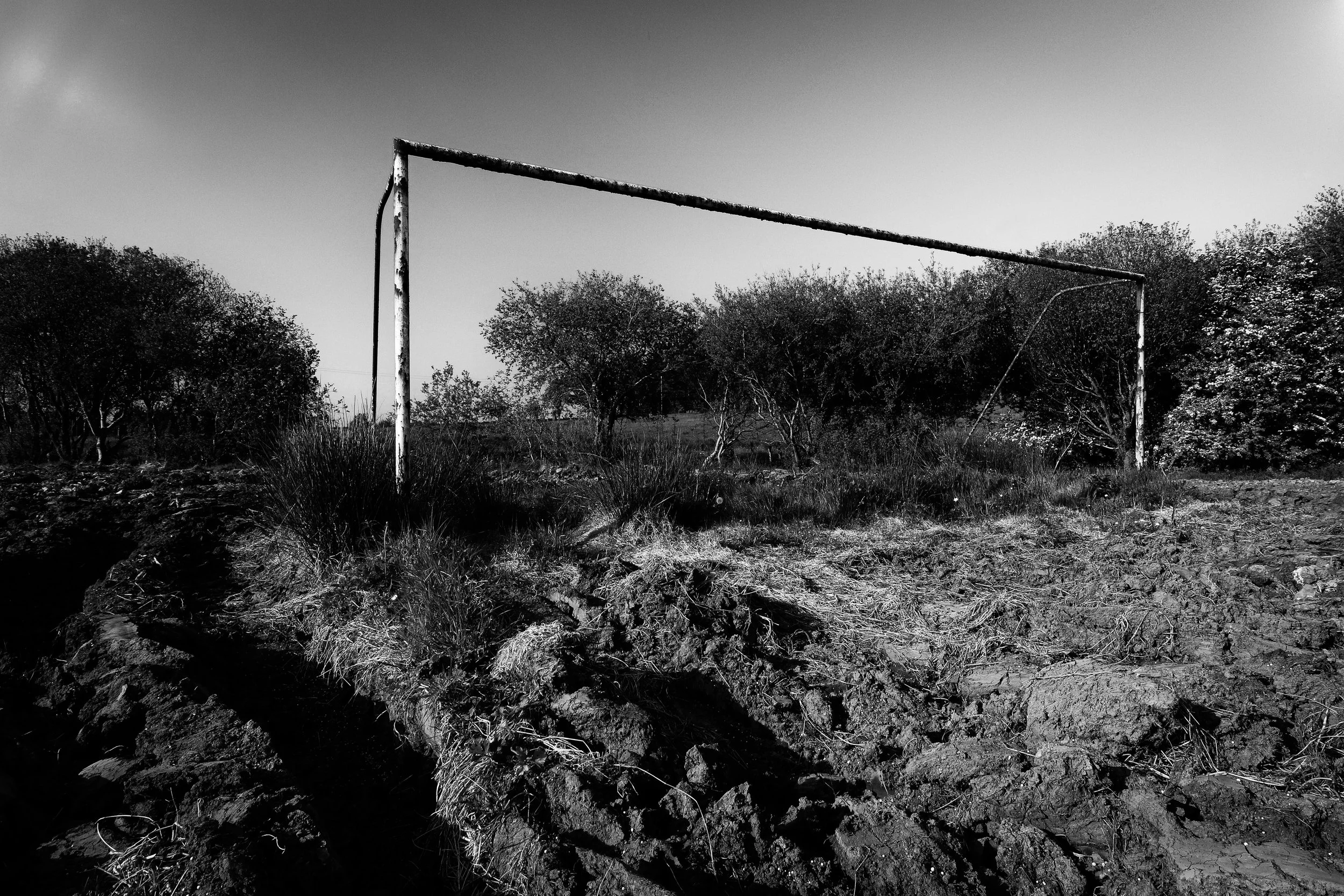 An old, rusted soccer goalpost in a field with dry grass, surrounded by trees, under a partly cloudy sky.