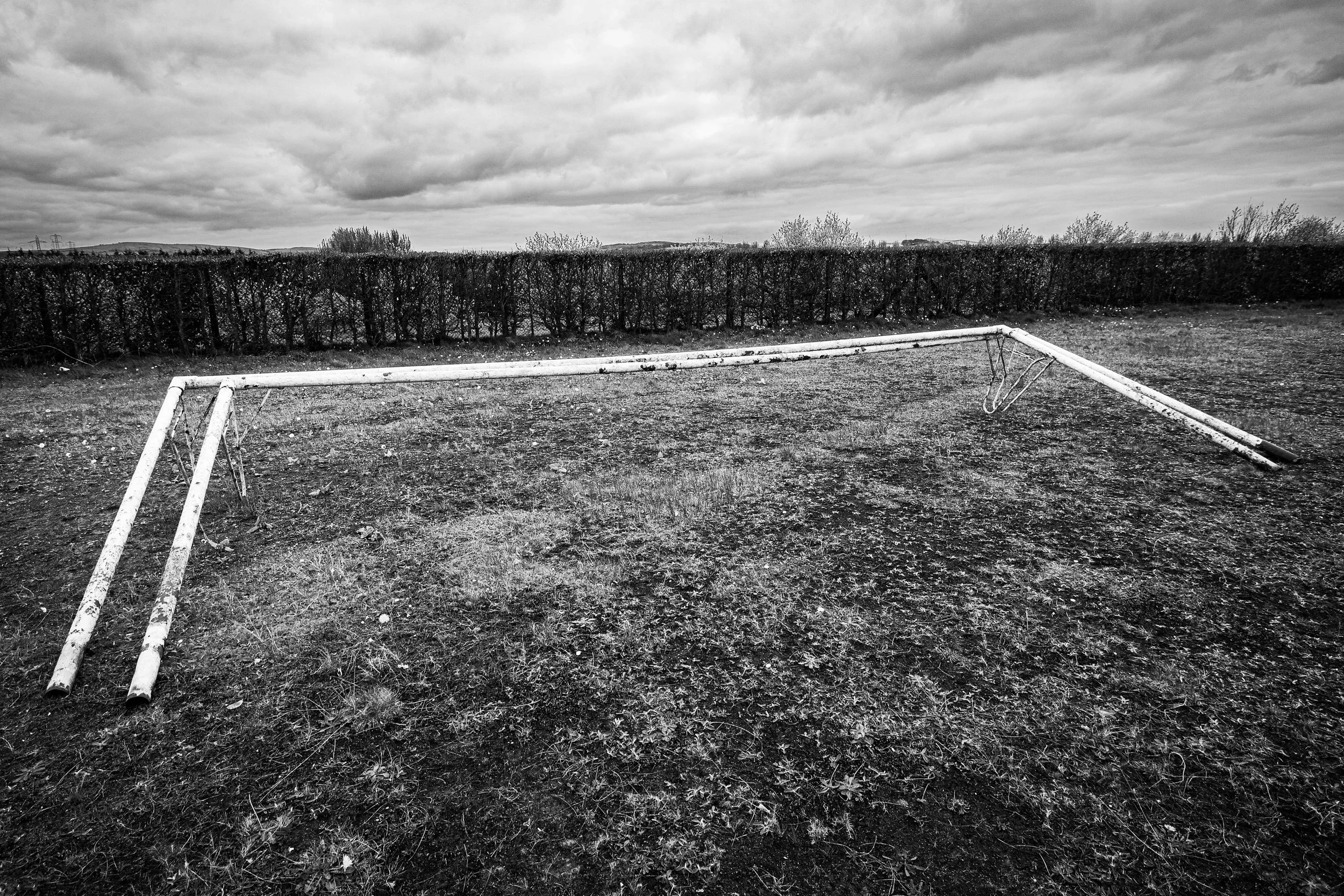 Empty weathered soccer goal on an empty field with a hedge and cloudy sky in the background.