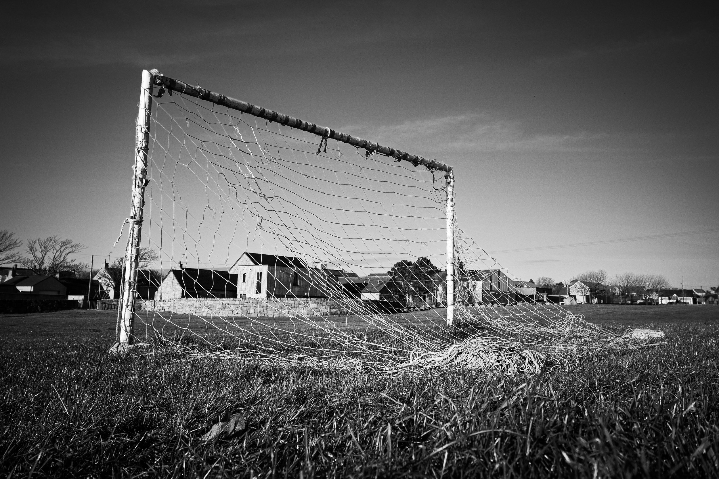 A worn soccer goal with a torn net on a grassy field, in front of a row of houses under a mild sky.