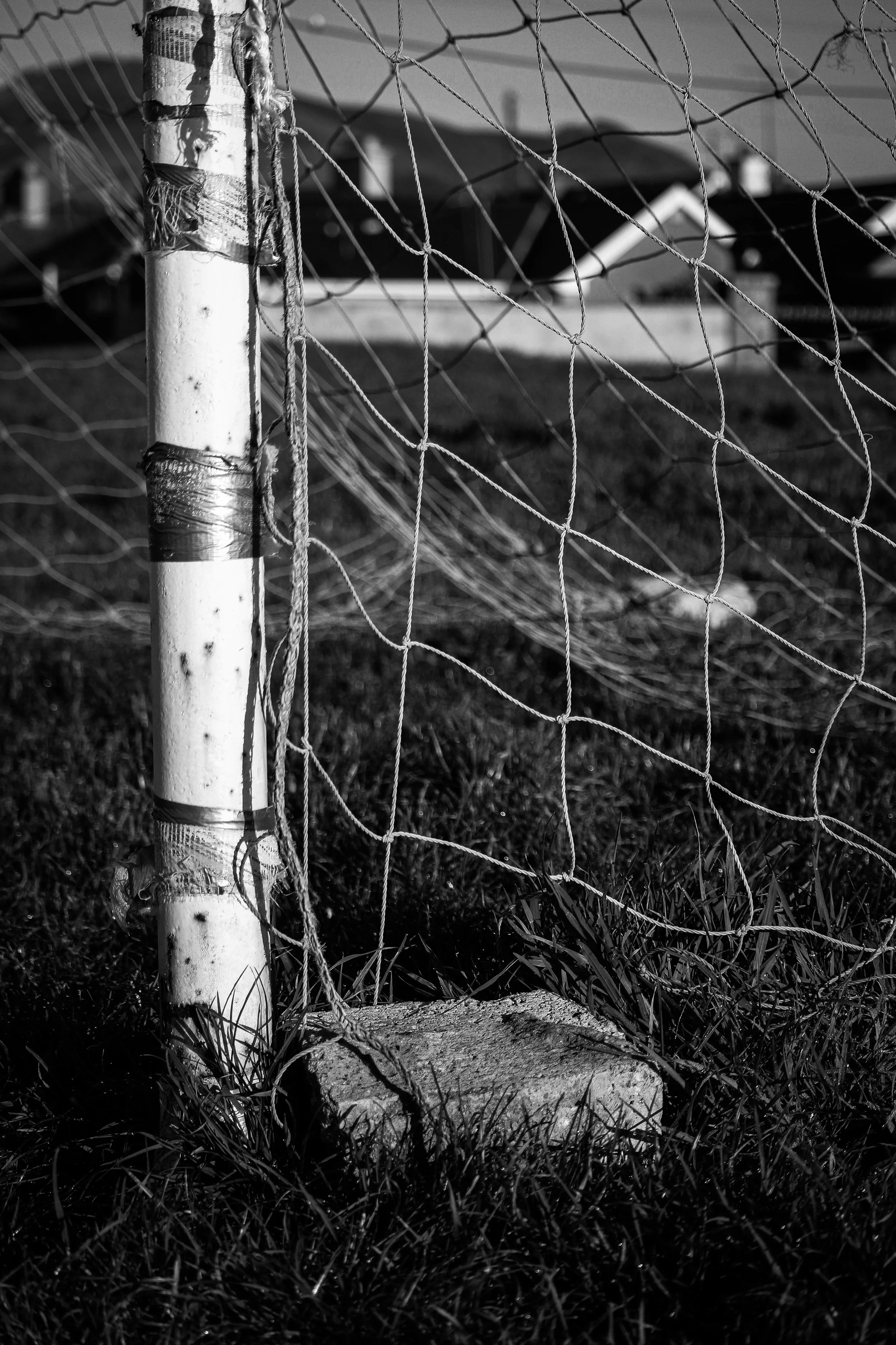 Close-up of a damaged soccer goal post and net in black and white, with a house in the background.