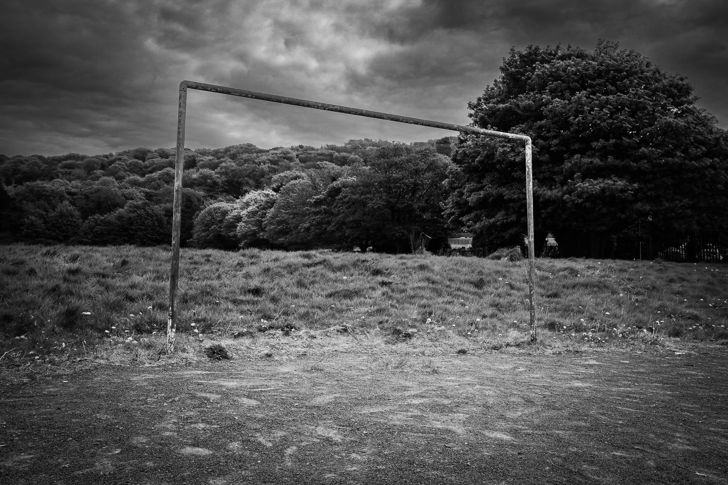 An empty soccer goal on a grassy field with trees and cloudy sky in the background, in black and white.