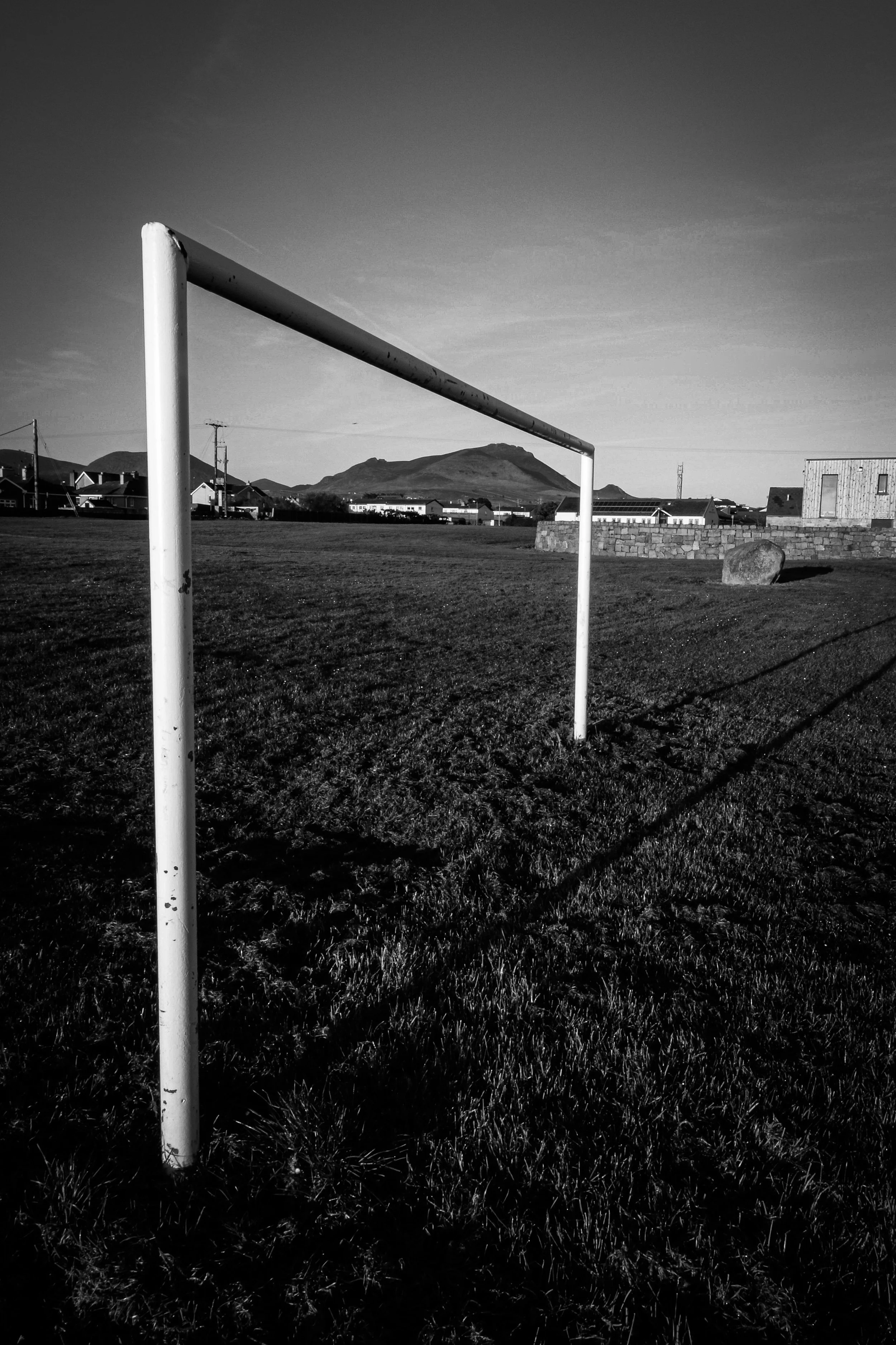 Black and white photo of a sports field with an empty goalpost in the foreground and mountains in the background.