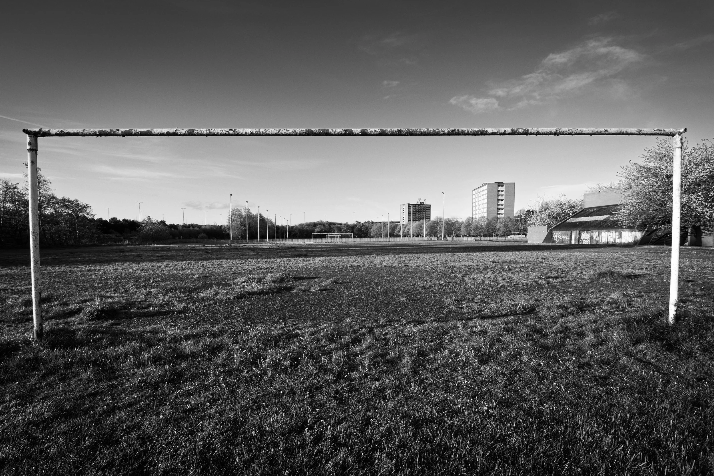 An empty soccer field with a goalpost in the foreground, surrounded by grass and distant buildings under a partly cloudy sky.