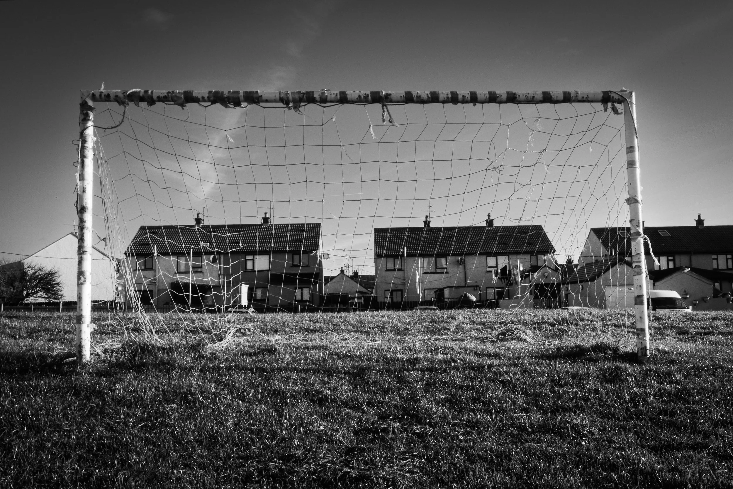 An old soccer goal net on a grassy field with residential houses in the background in black and white.