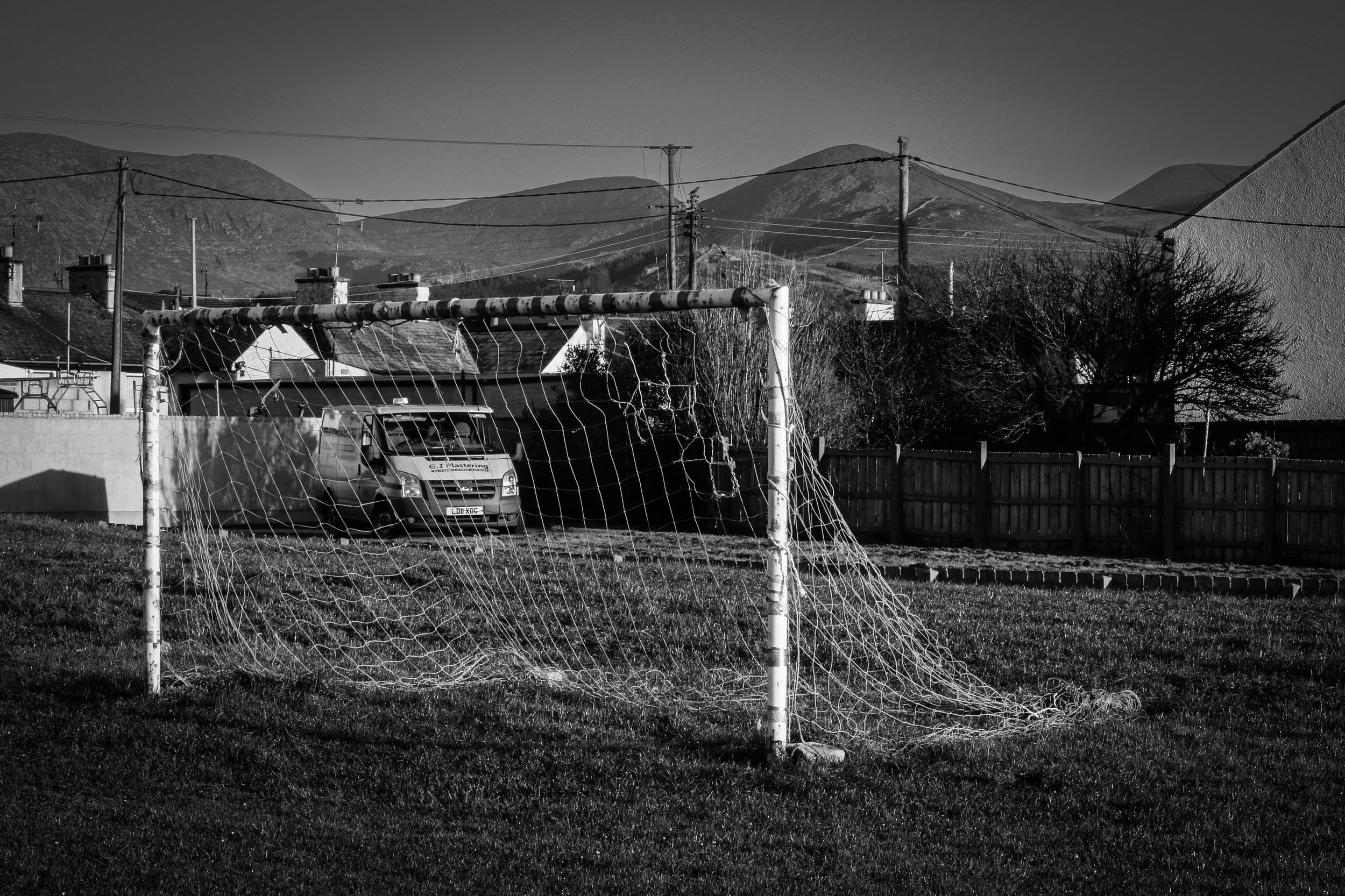 An old, damaged soccer goal with a frayed net on a grassy field, with vehicles and houses in the background, and mountains in the distance.
