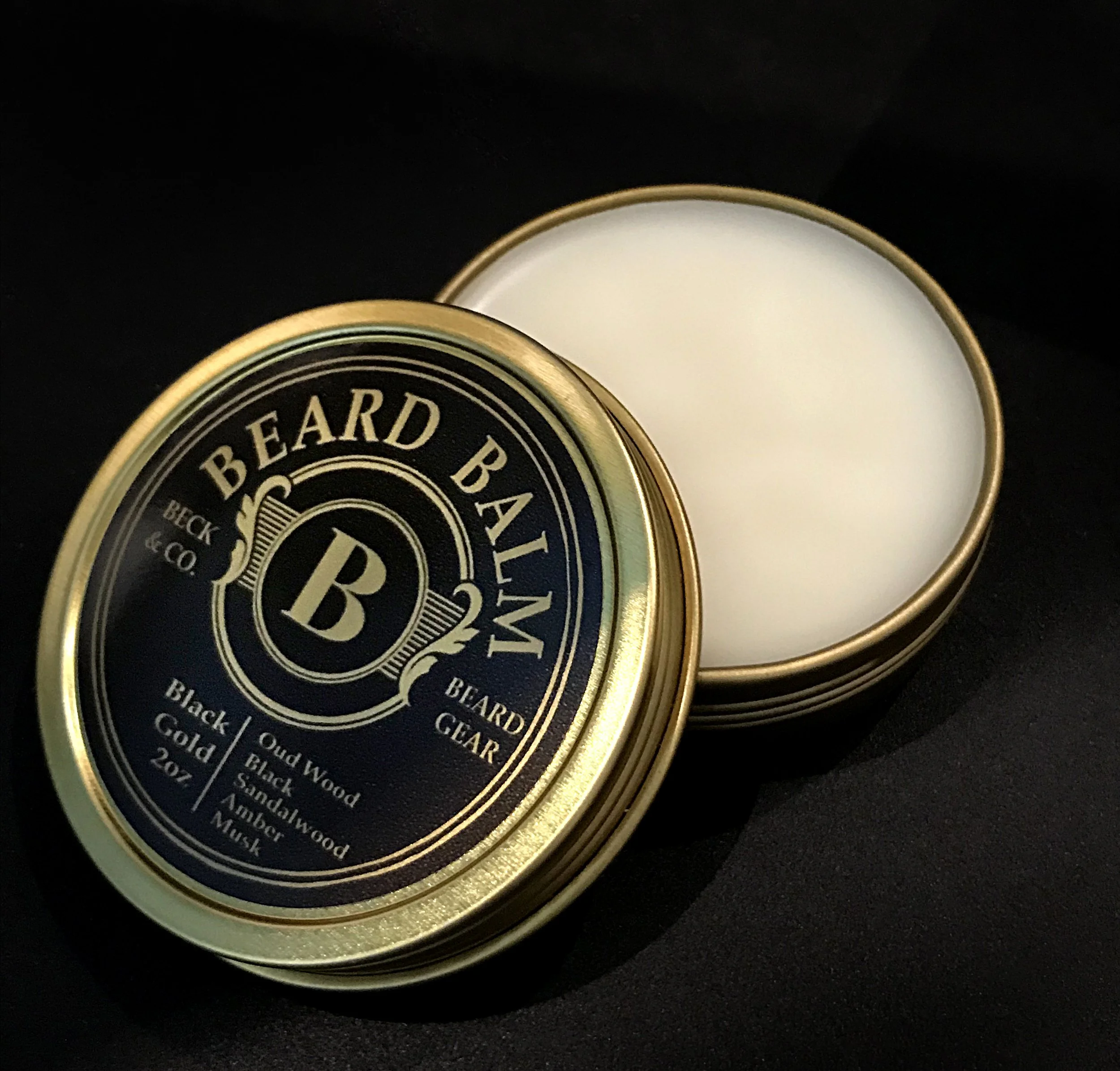 A round, gold tin of beard balm with a black and gold label, open to reveal white balm inside.