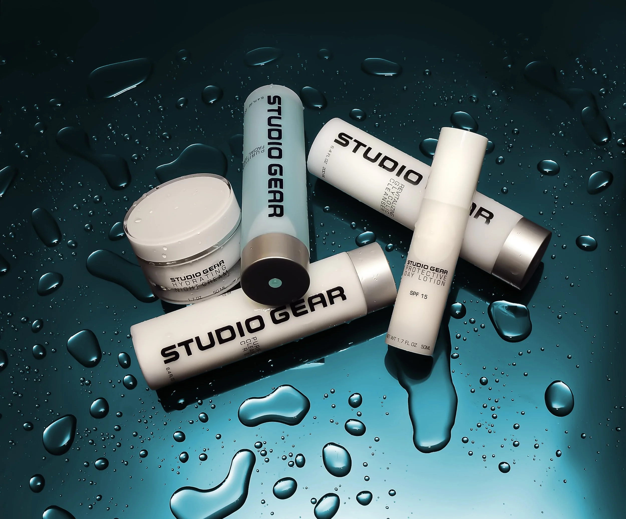 Studio Gear skincare products arranged on a dark, wet surface with water droplets, including creams and lotions in white containers with black labels.