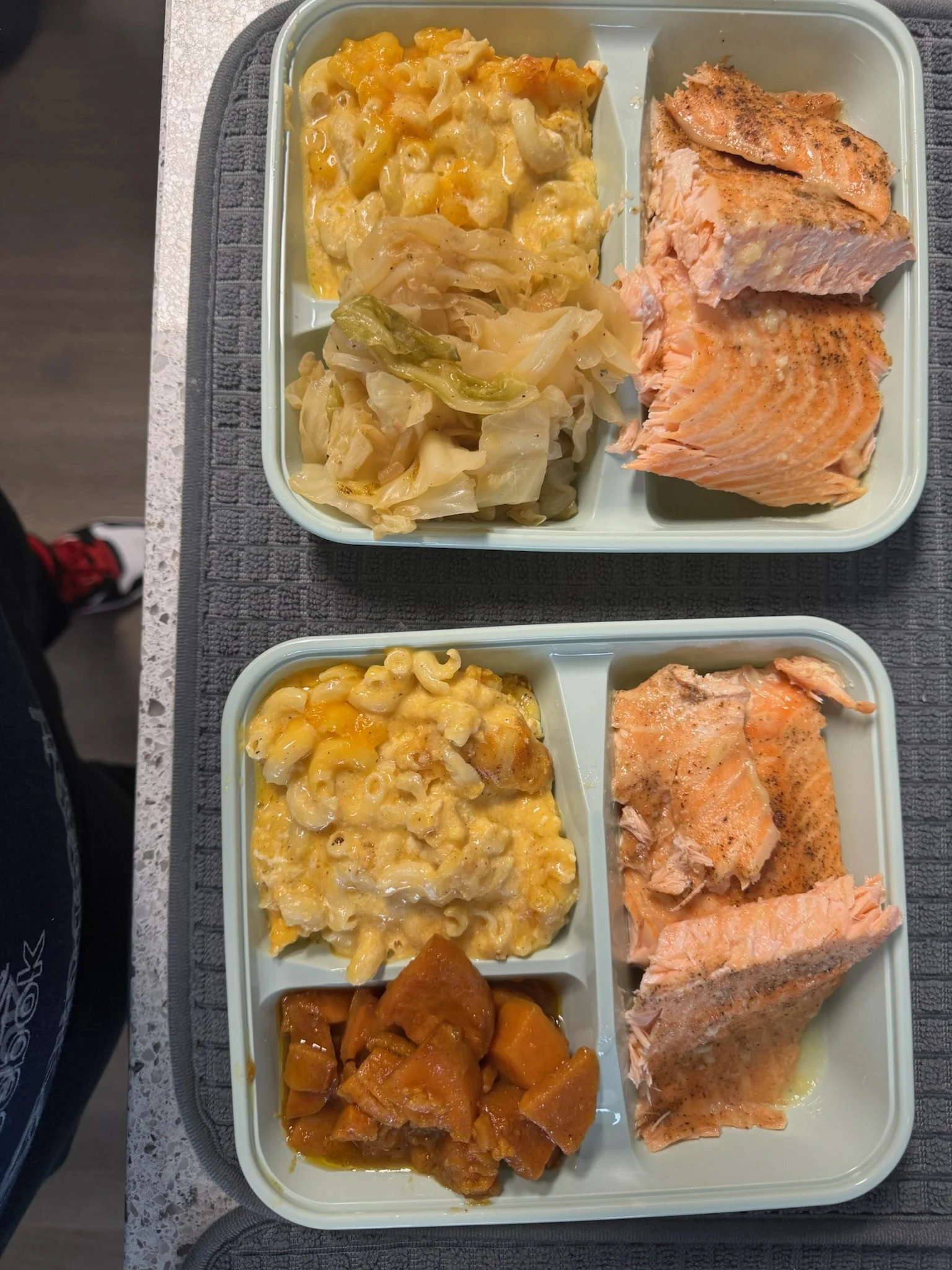 Two cafeteria trays, each with sections containing macaroni and cheese, cooked cabbage, baked salmon, and sweet potatoes.