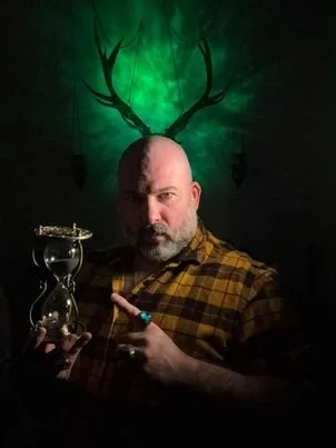 A man with a beard holding an hourglass in one hand and a glass of wine in the other, with green glowing antlers projected on the wall behind him.