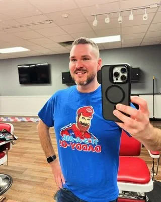 A smiling man taking a selfie in a room with a mirror. He is wearing a blue T-shirt with a Santa Claus graphic and the text "Merry Christmas Daddy". The room has red chairs and a television mounted on the wall.
