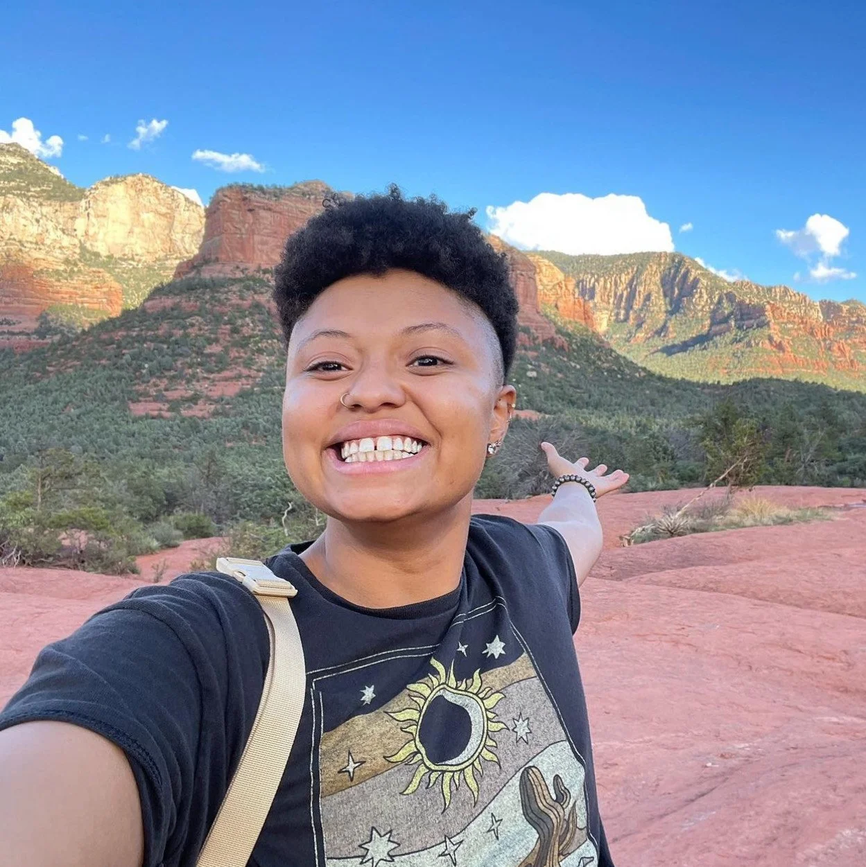 A smiling person with short curly hair and a nose ring posing in front of red rock formations and green mountains under a blue sky with clouds, pointing towards the landscape.