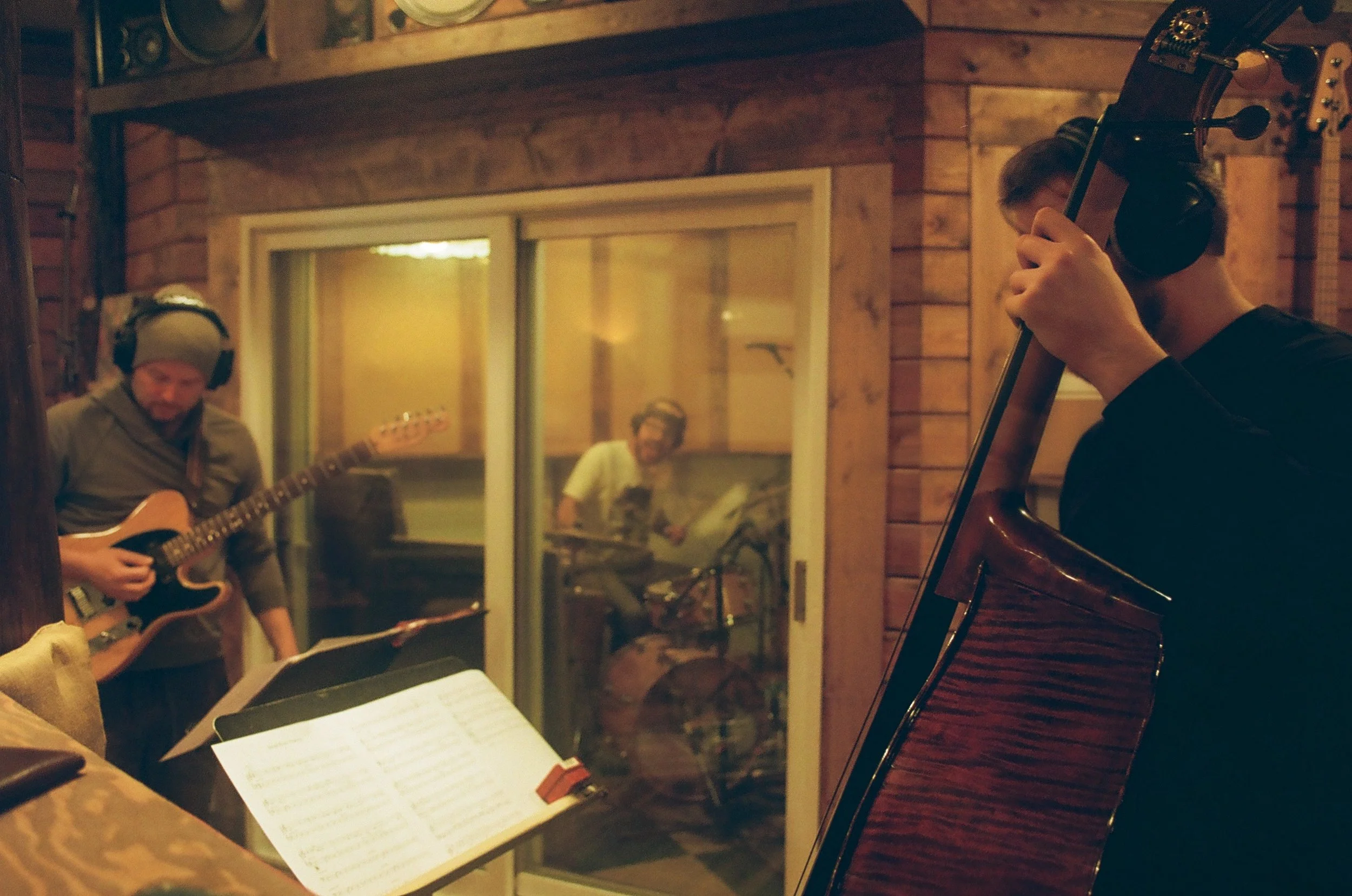 Musicians recording in at Holy Cow studio; one playing guitar, another playing double bass, and a third playing drums in a separate booth. Music sheets are visible.