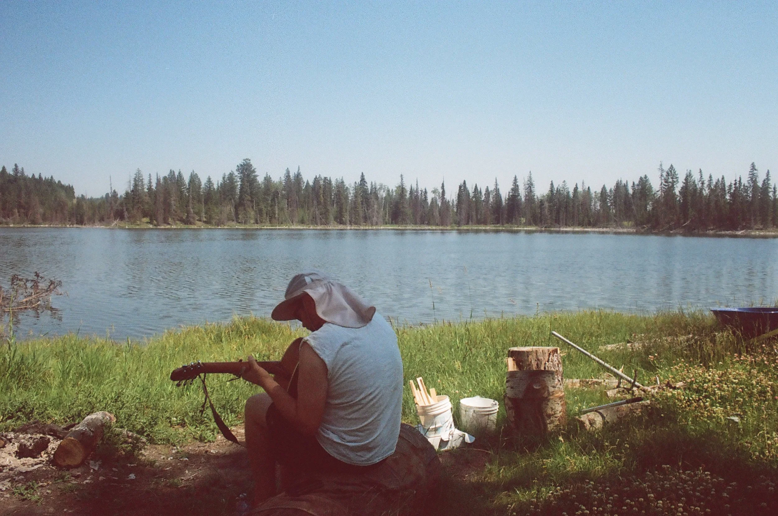 Person sitting by a lakeside playing a guitar, with a sun hat, surrounded by nature, visible tree line across the water.