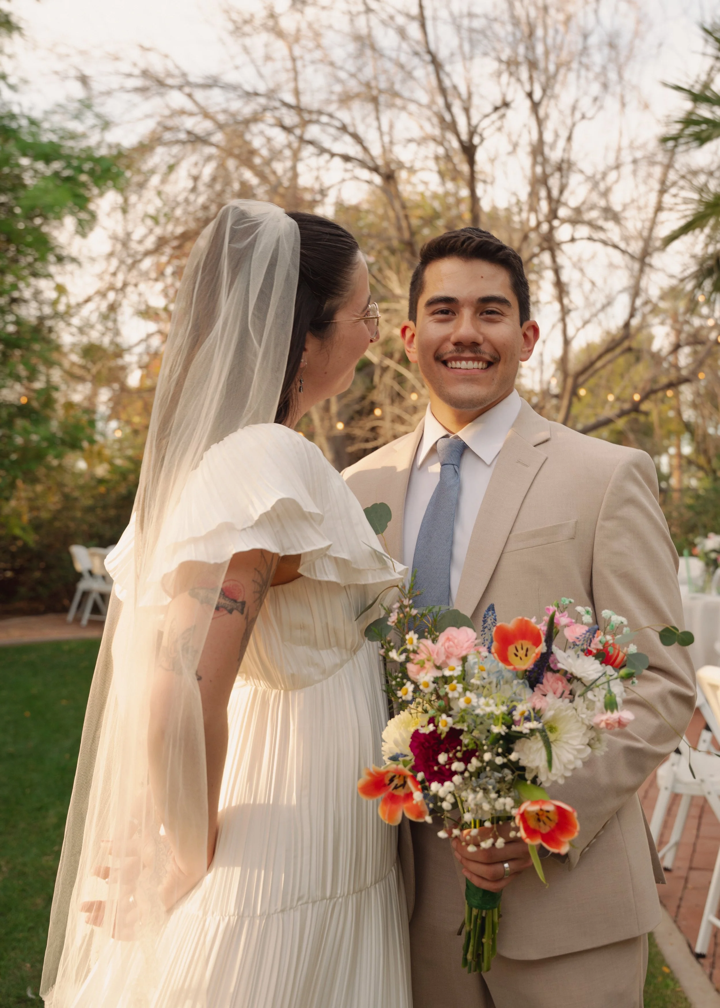 A smiling couple dressed in wedding attire, outdoors with trees in the background, holding a bouquet of colorful flowers.