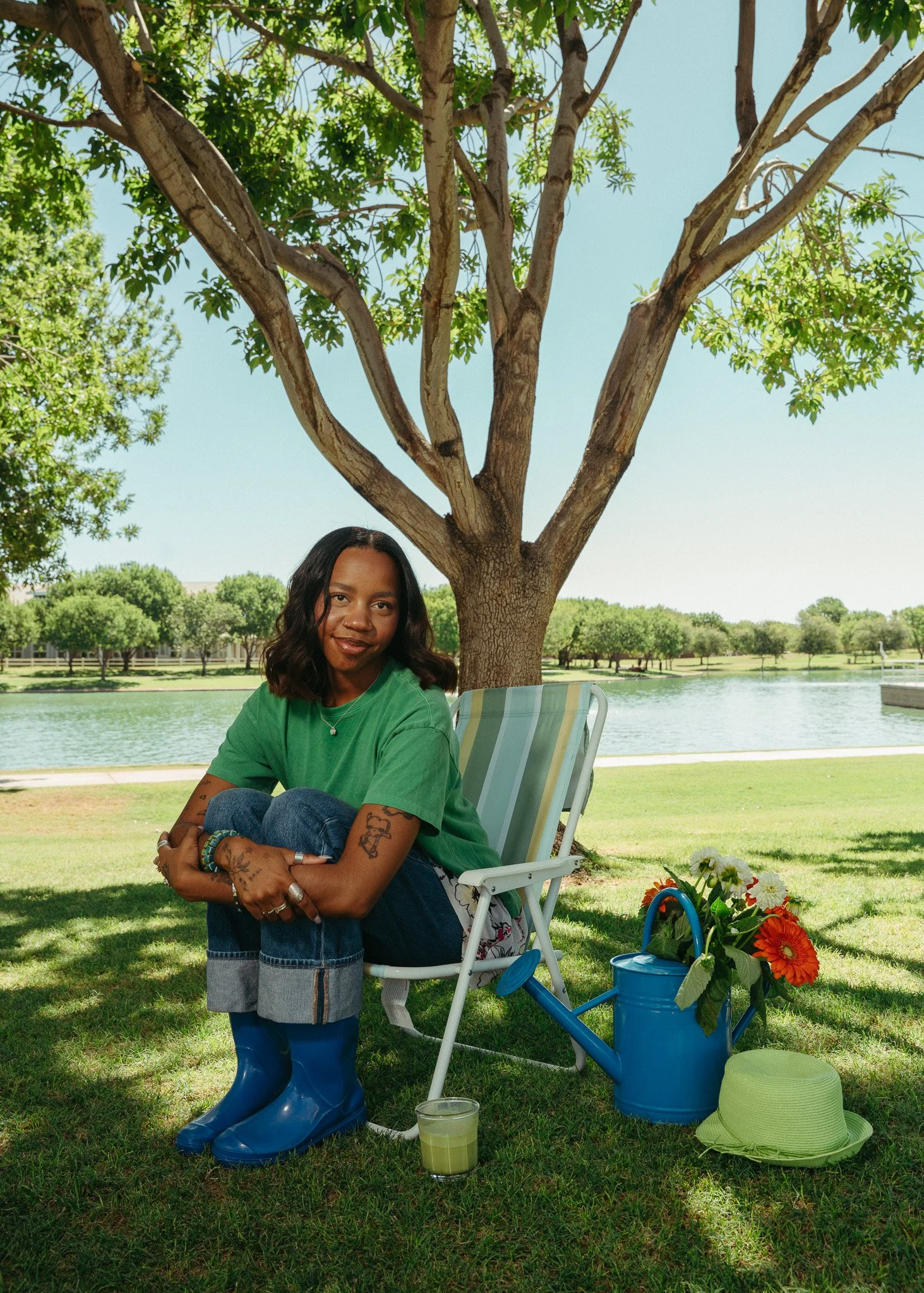 A woman with tattoos in a green t-shirt sitting with her knees up on a lawn chair outdoors. Behind her is a tree with green leaves and a pond in the background. On the grass in front of her are a yellow-green hat, a small potted plant with orange and white flowers, a blue watering can, a glass of green drink, and a green cushion.