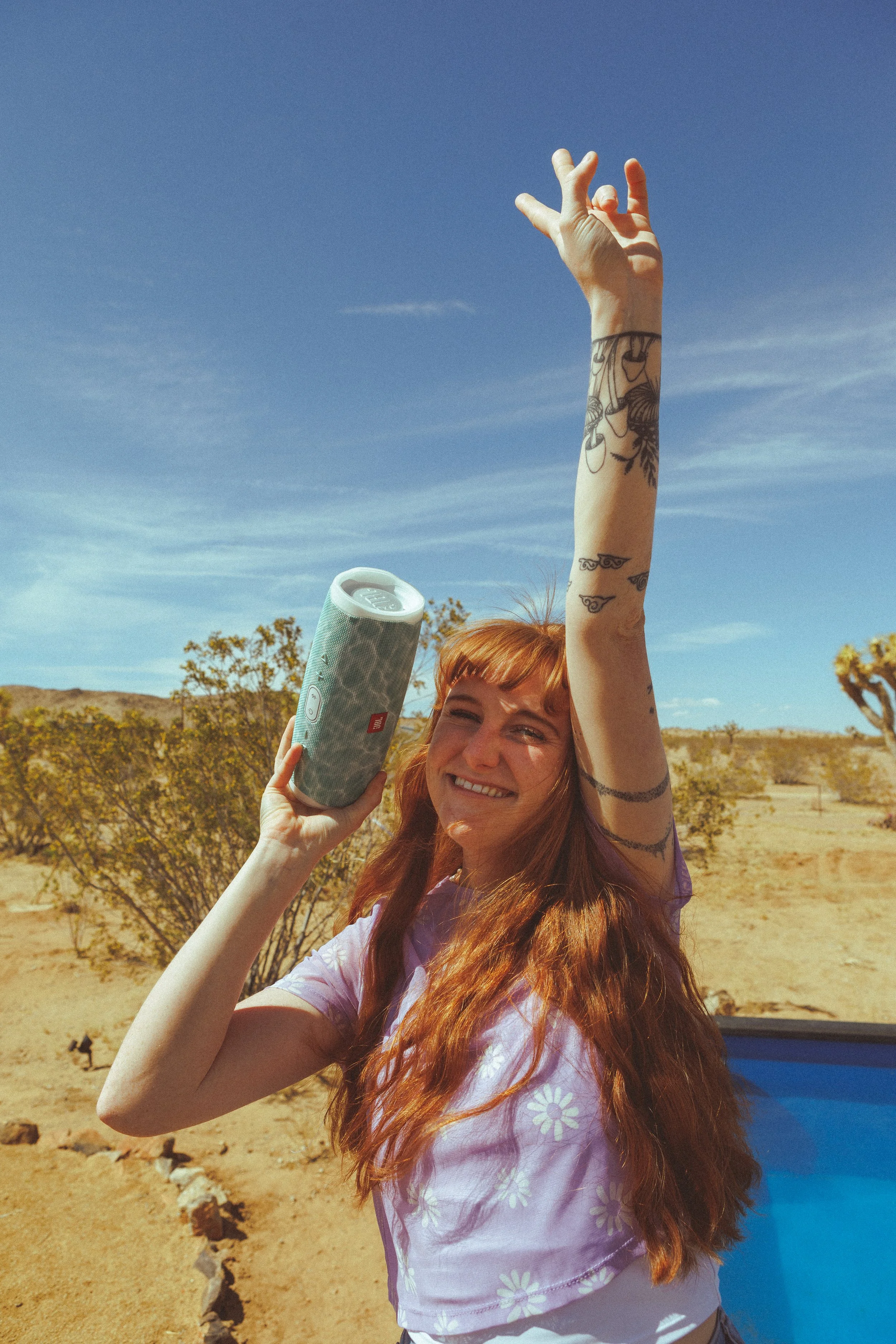 A young woman with red hair and tattoos on her arm is outdoors in a desert landscape, smiling and holding a portable speaker in one hand and raising her other hand in the air under a blue sky.