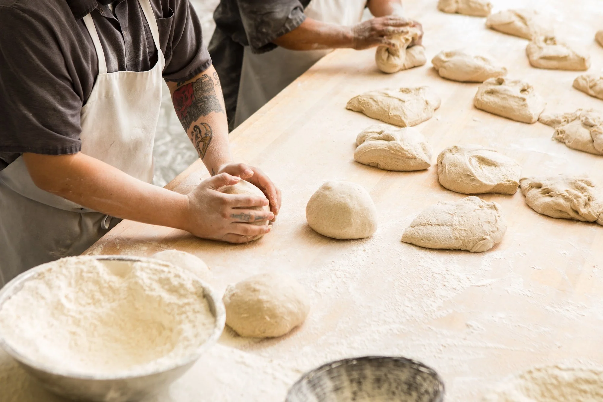 Two bakers hand-shaping rounds of sourdough dough on a floured worktable at Grand Central Bakery, part of the scratch-made process behind every wholesale order