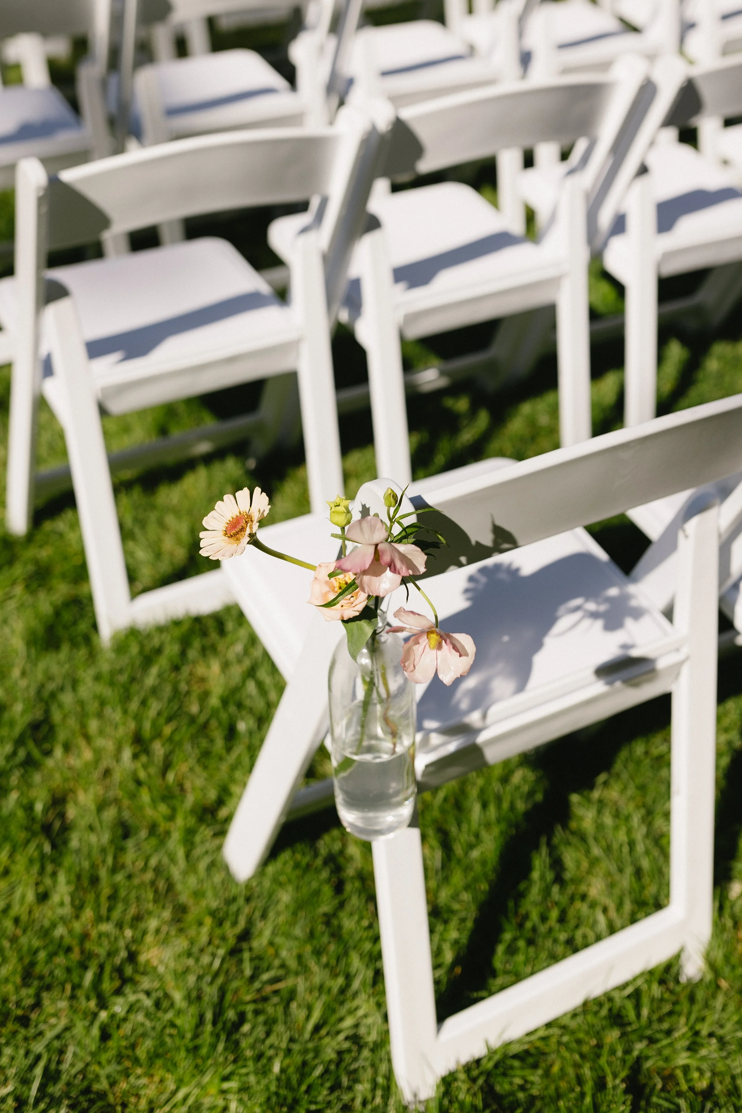White folding chairs arranged outdoors on grass with a small glass vase holding pink and white flowers attached to one of the chairs.