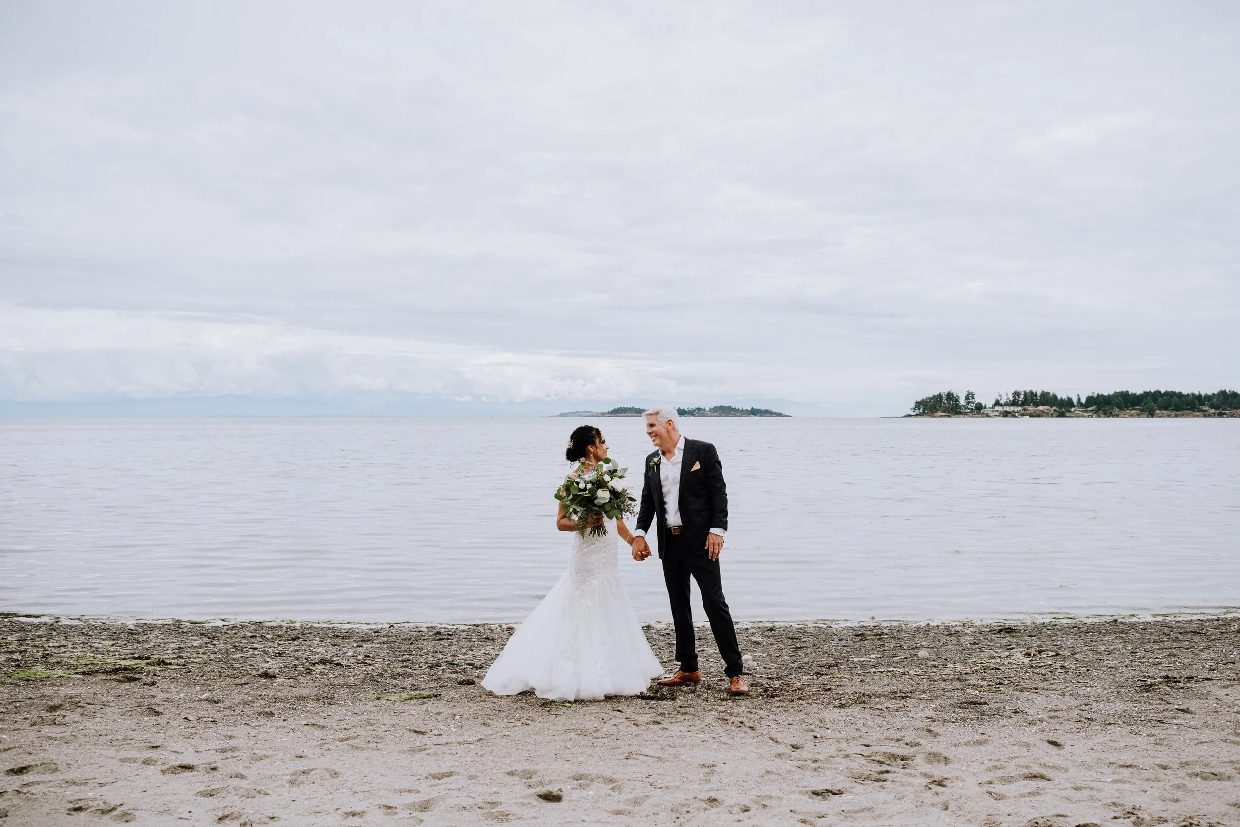Bride and groom holding hands and smiling at each other on a beach with water and small islands in the background during their wedding.