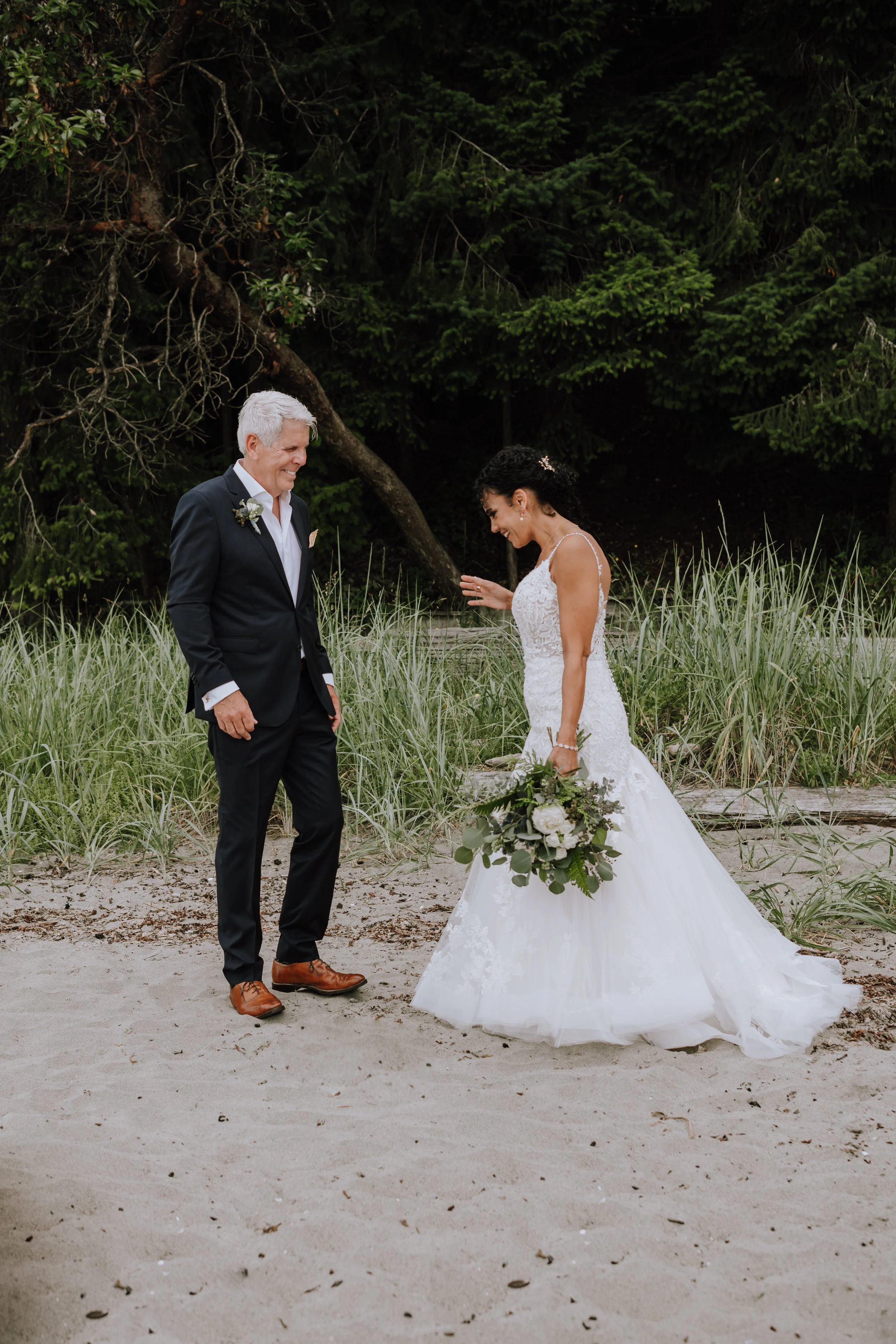 Bride and groom on a beach, smiling and sharing a moment during their wedding, with trees and grass in the background.