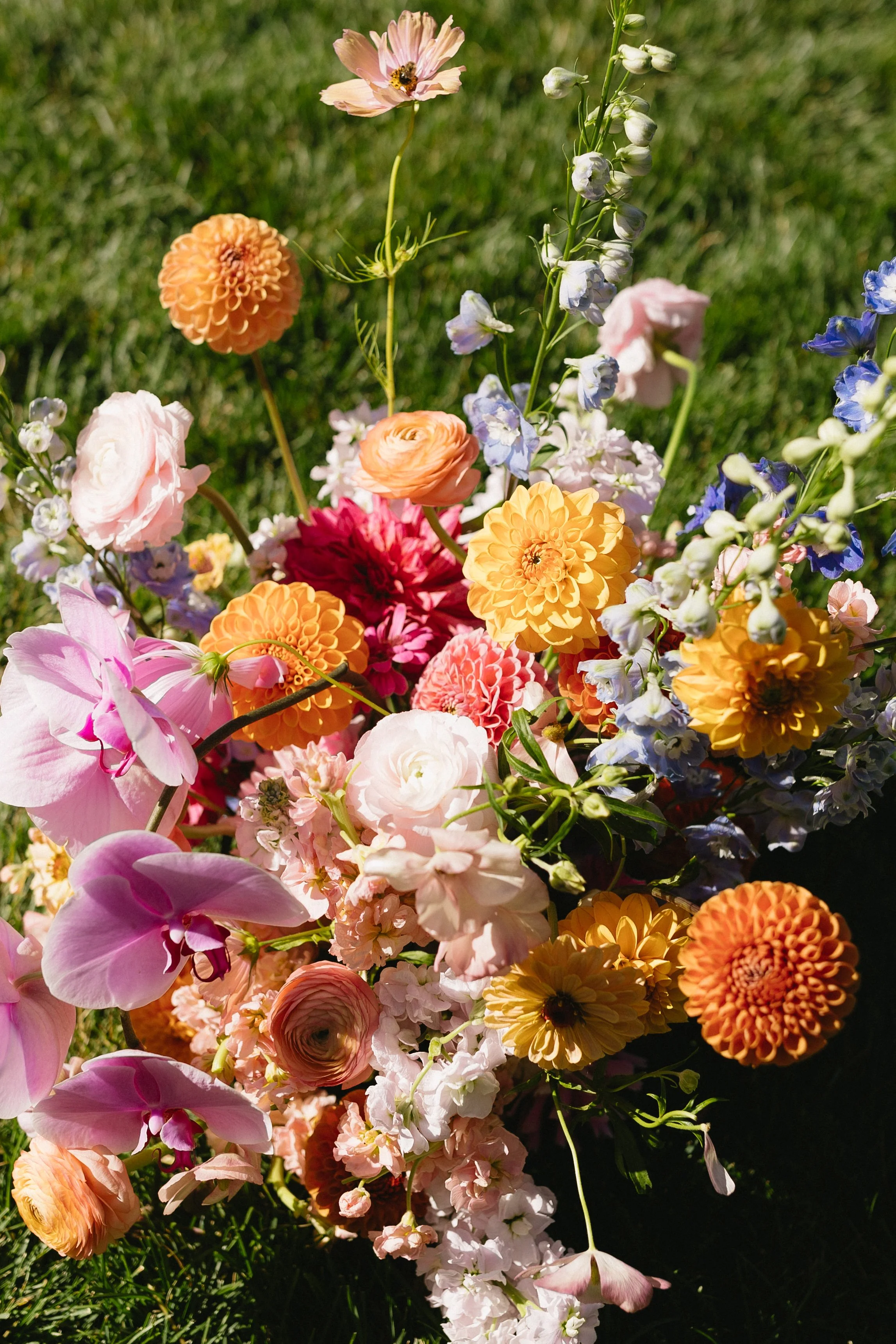Colorful bouquet of various flowers including dahlias, roses, orchids, and delphiniums on green grass.