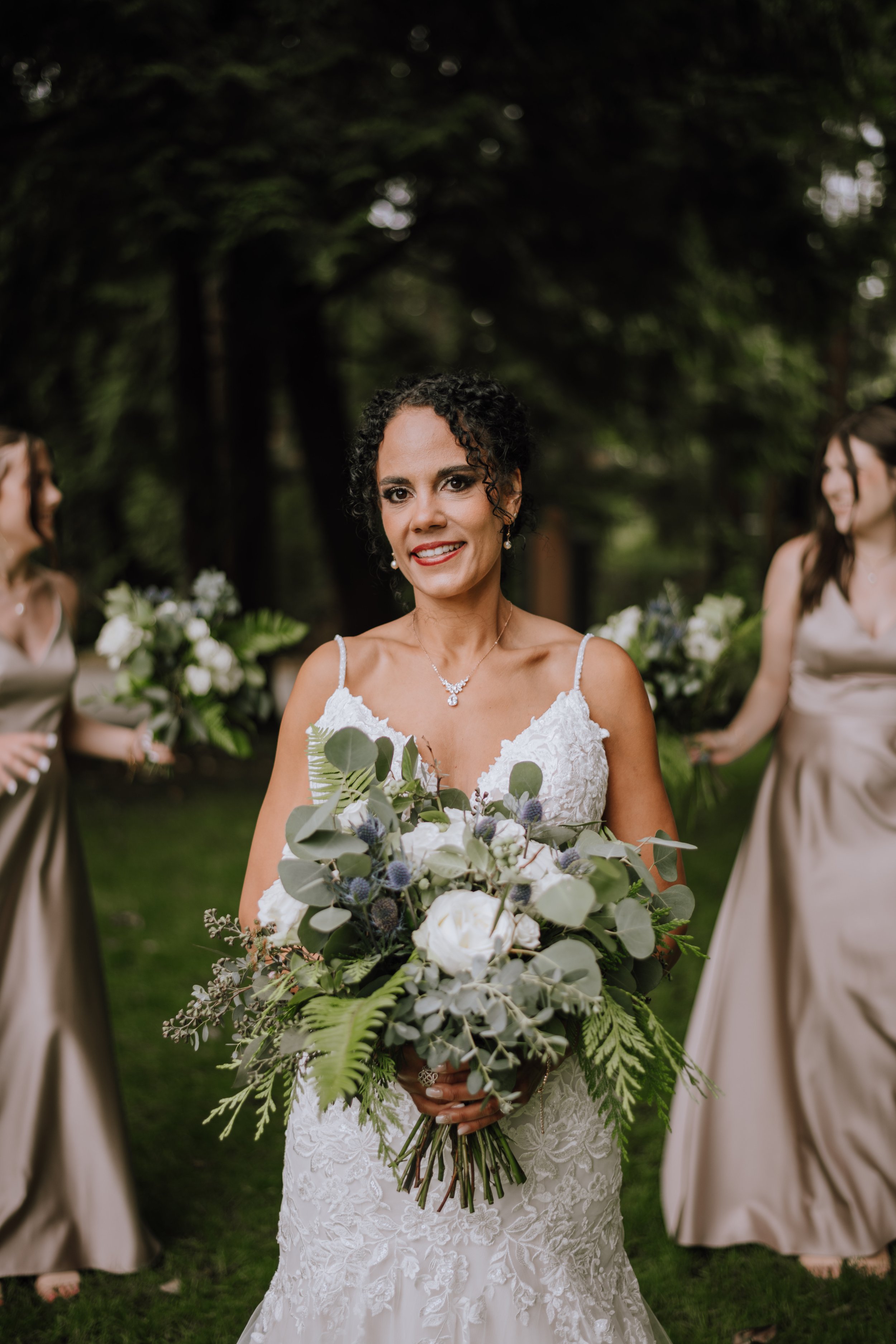 A bride in a white lace wedding dress holding a bouquet of white roses, greenery, and thistle, smiling outdoors with two bridesmaids holding matching bouquets in the background.