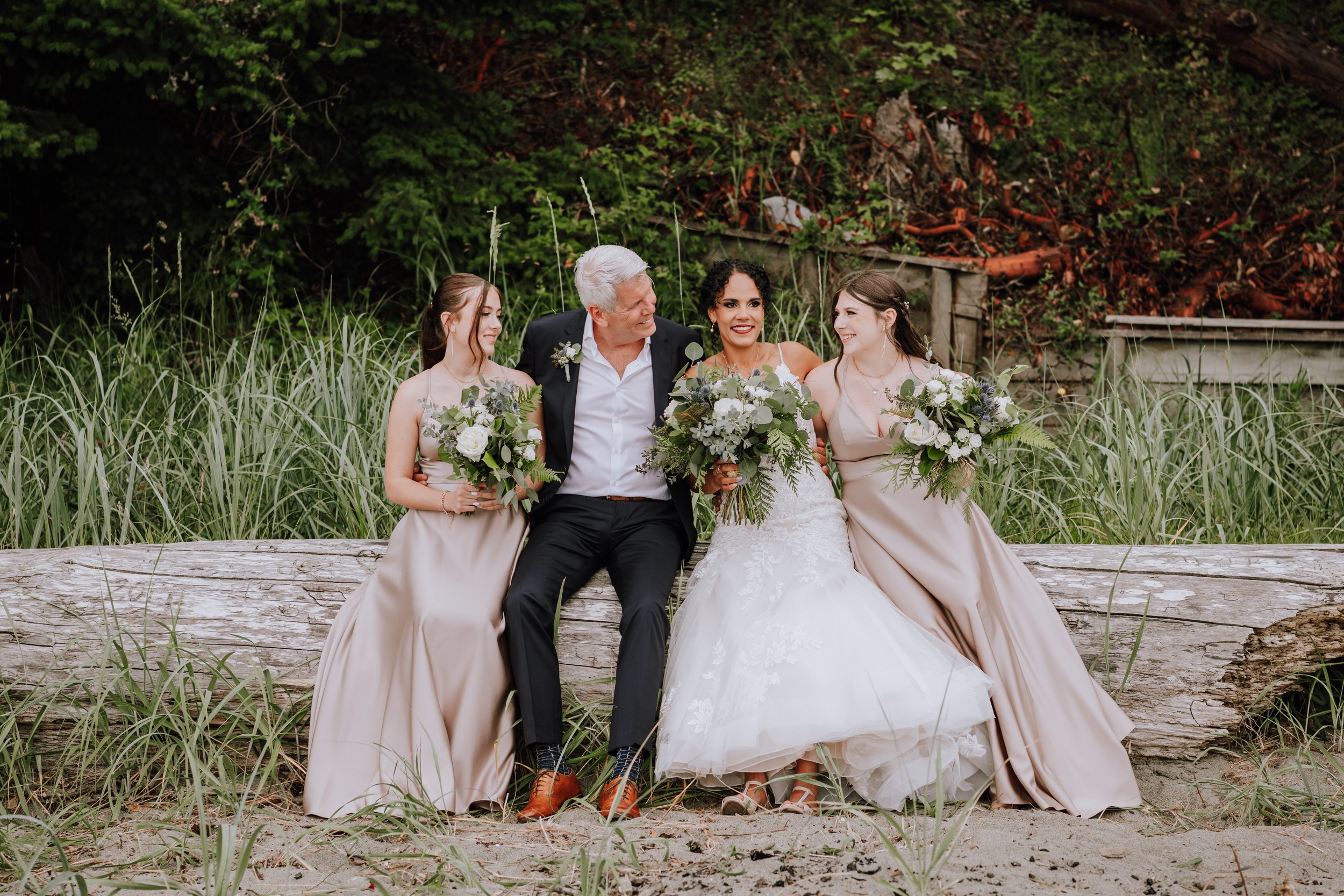A group of five people sitting on a log outdoors, including a bride in a white wedding dress, two bridesmaids in beige dresses, and two men, one in a black suit and the other in a dark blazer, all holding bouquets of flowers. They are smiling and loo