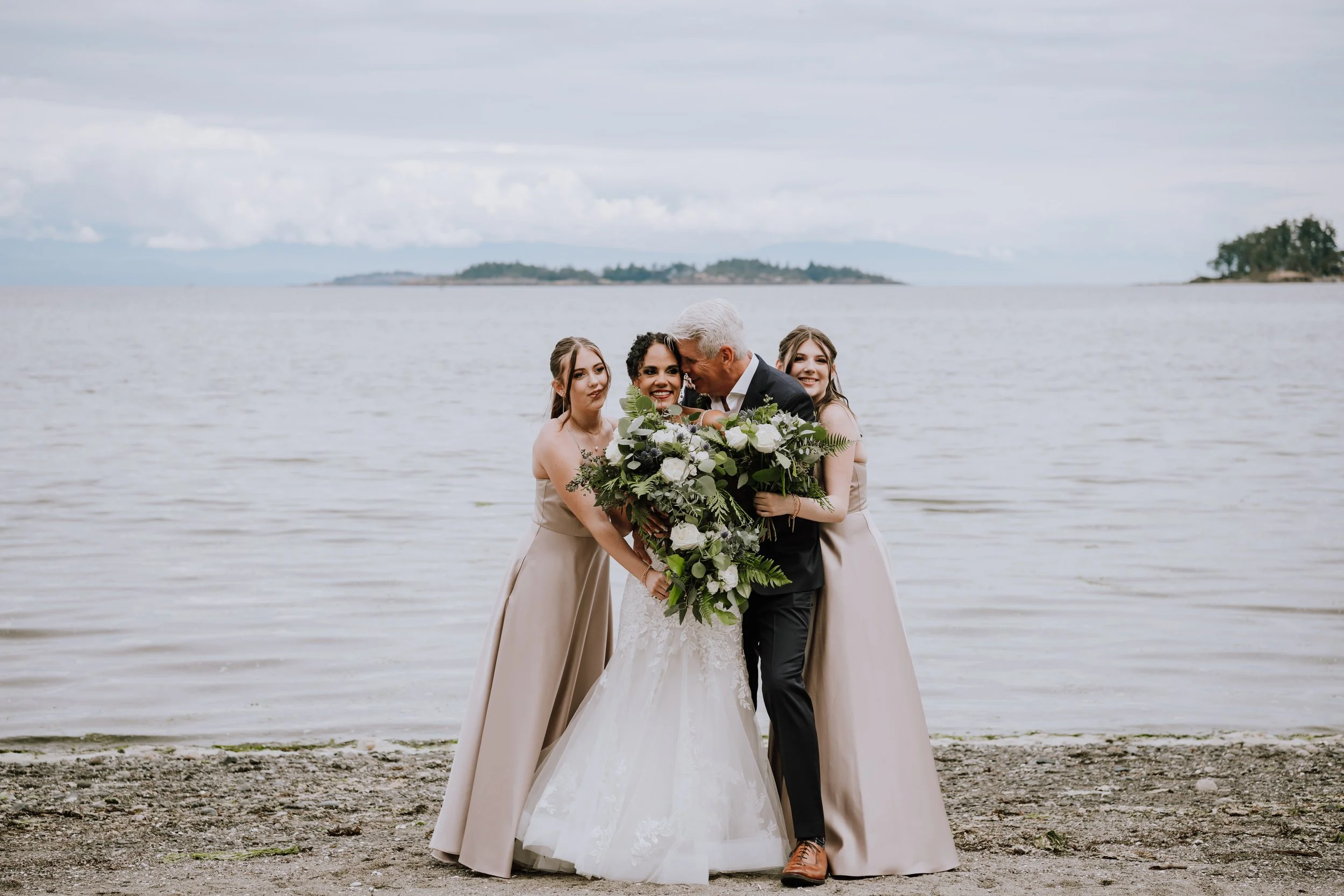 A wedding party by the water, with a bride, groom, and two bridesmaids holding bouquets, posing on a rocky beach under a cloudy sky.