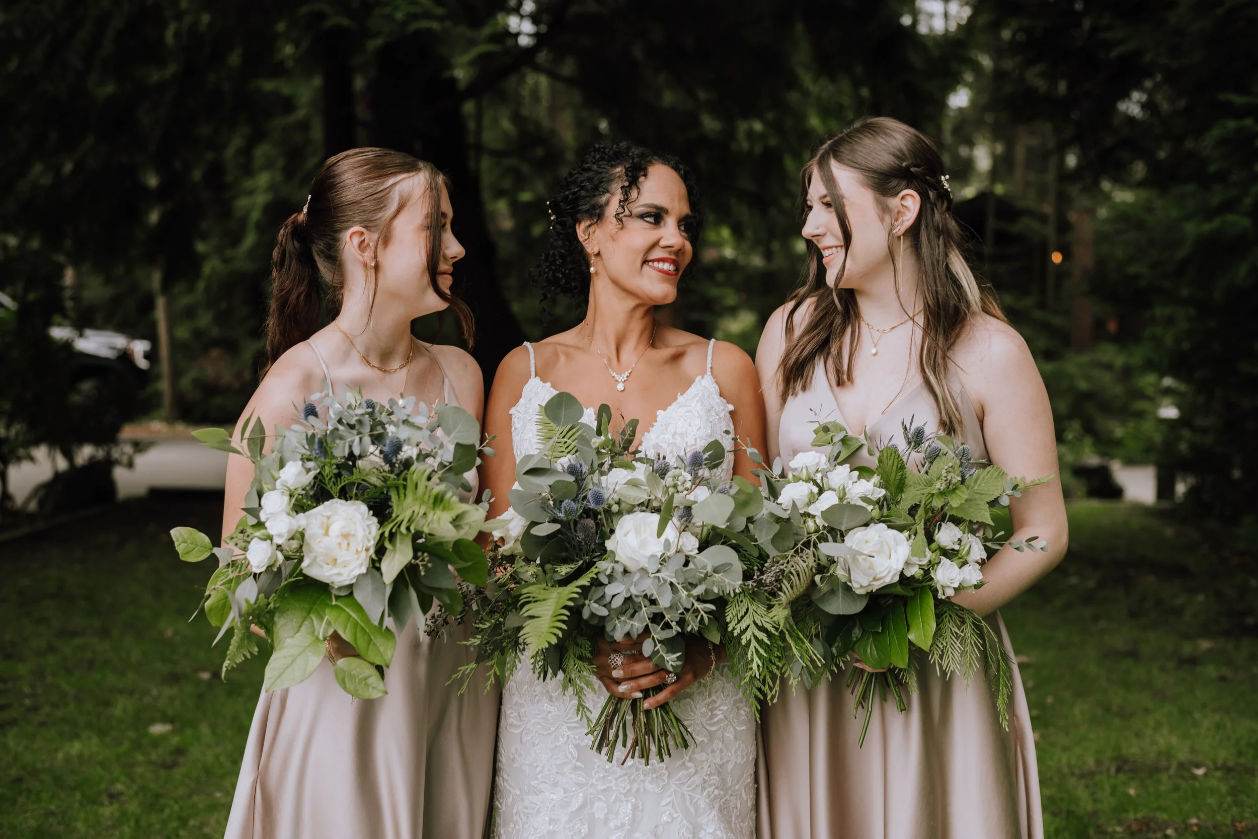 A bride with two bridesmaids holding bouquets of white flowers and greenery, standing outdoors in a wooded area.