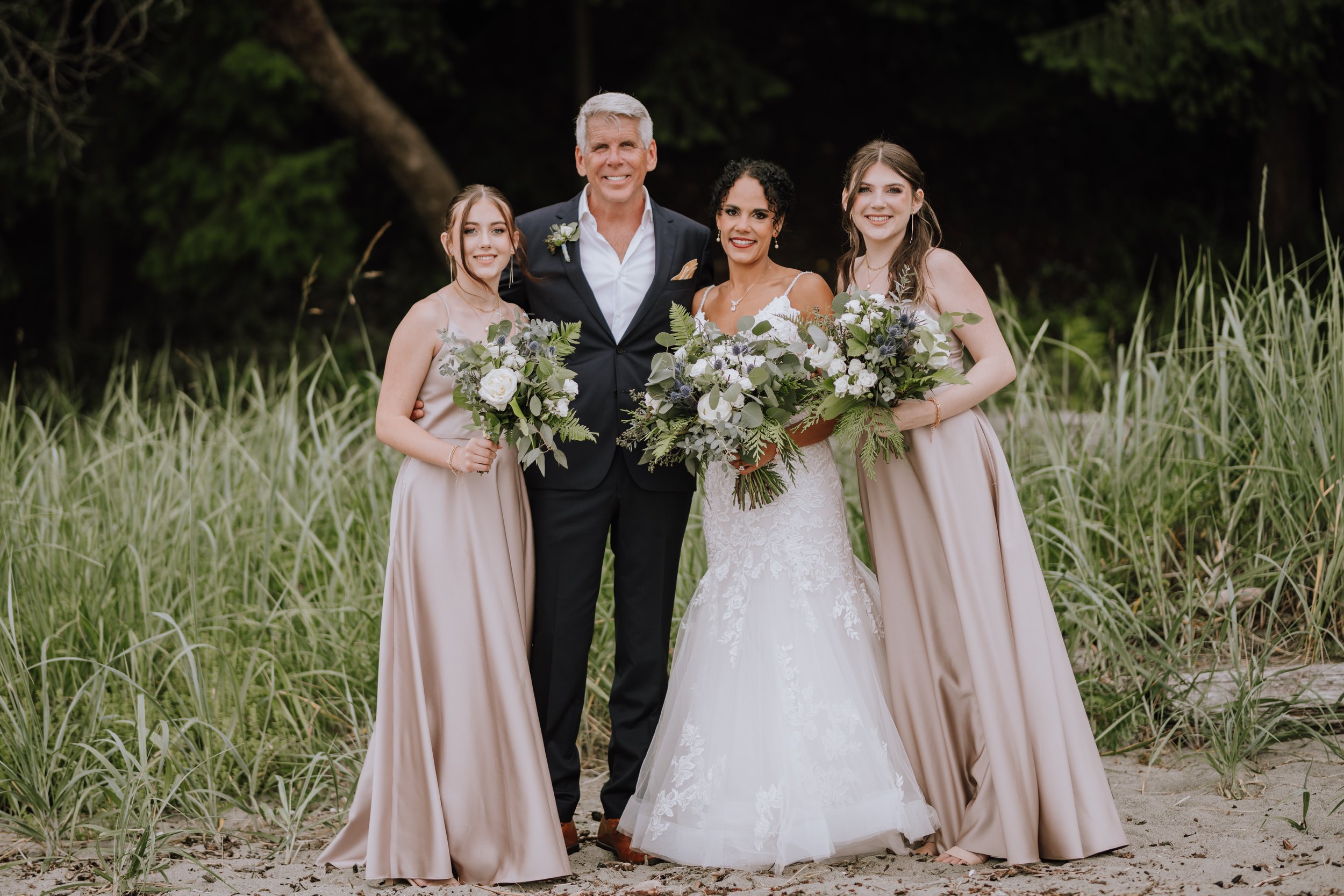 A bride in a white lace wedding gown standing with three women in beige dresses and a man in a suit, all smiling, outdoors in a grassy area with trees in the background, holding bouquets of white and green flowers.