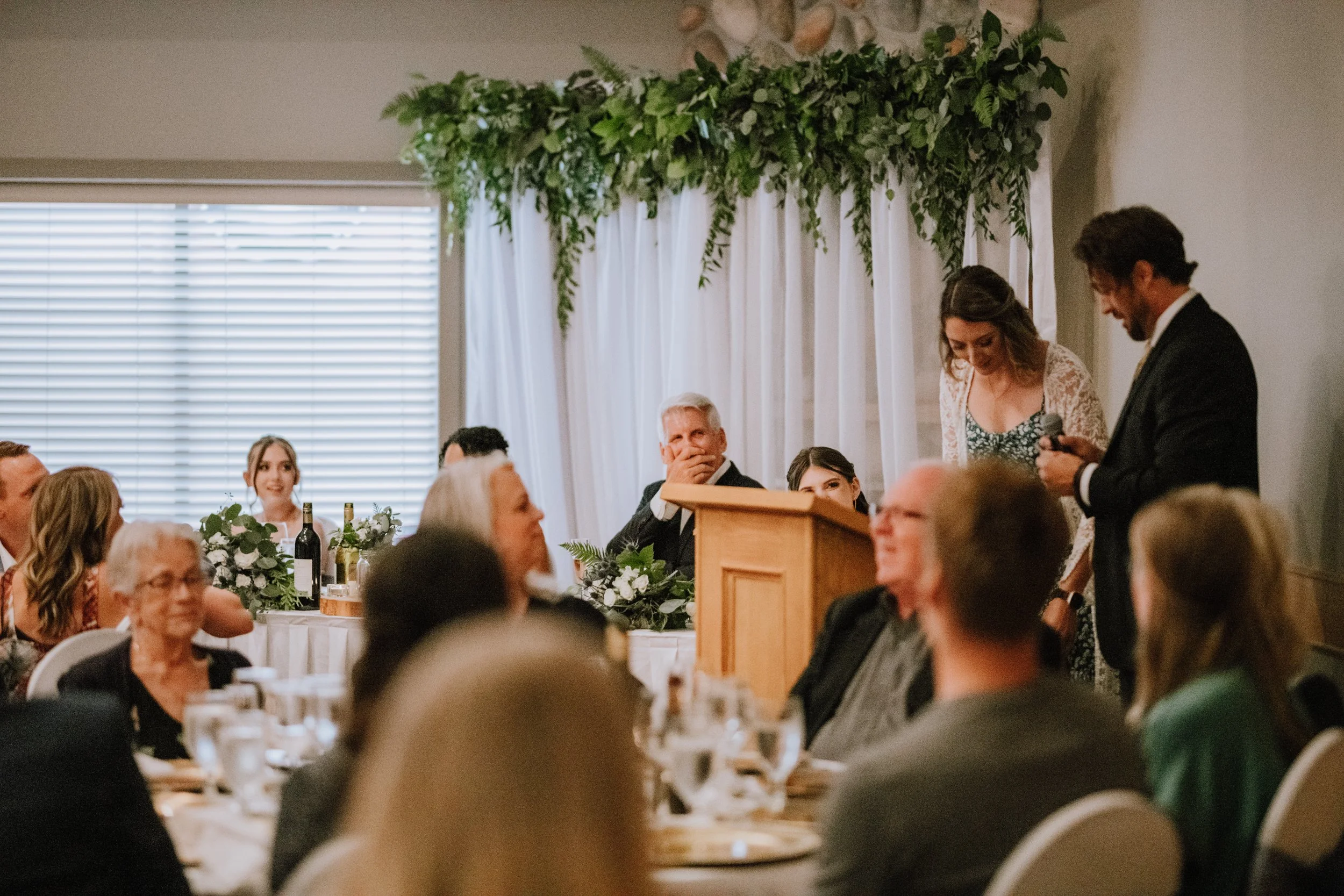 A wedding reception where a woman is giving a speech at a podium, with guests seated at tables, some looking toward her and others smiling, decorated with greenery and wine bottles.