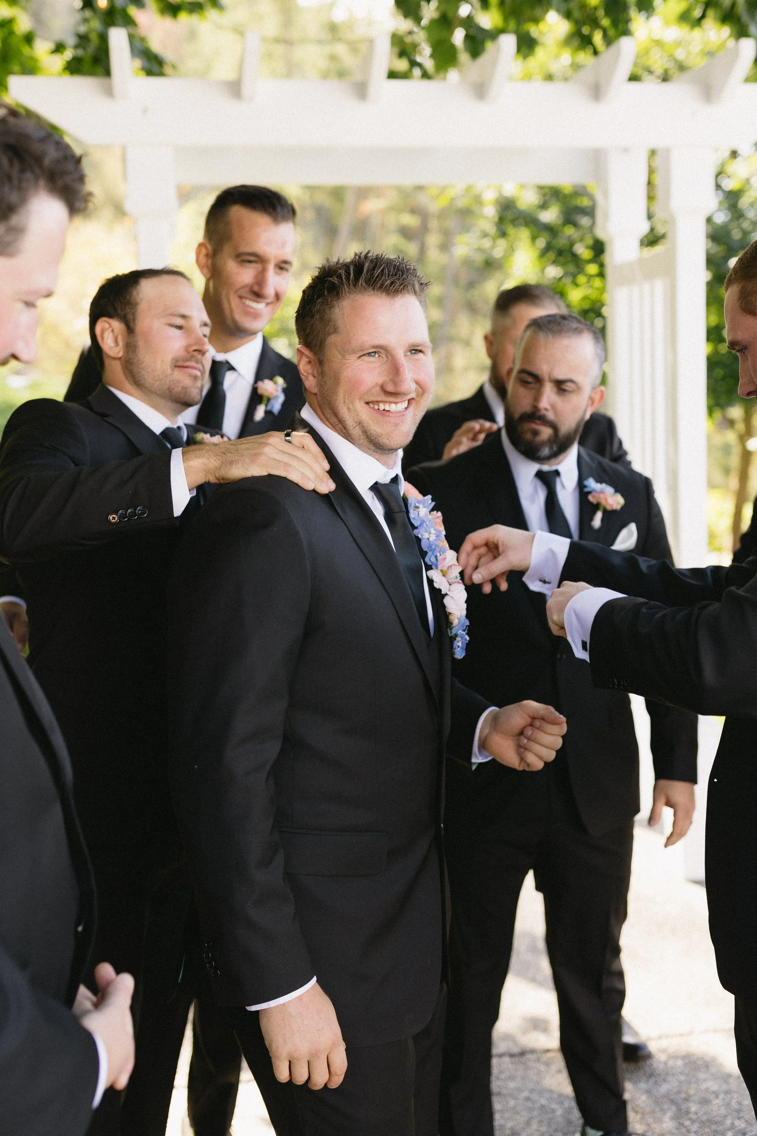 Groom in black tuxedo smiling at wedding ceremony, surrounded by groomsmen also in tuxedos with boutonnières, outdoors with greenery in background.