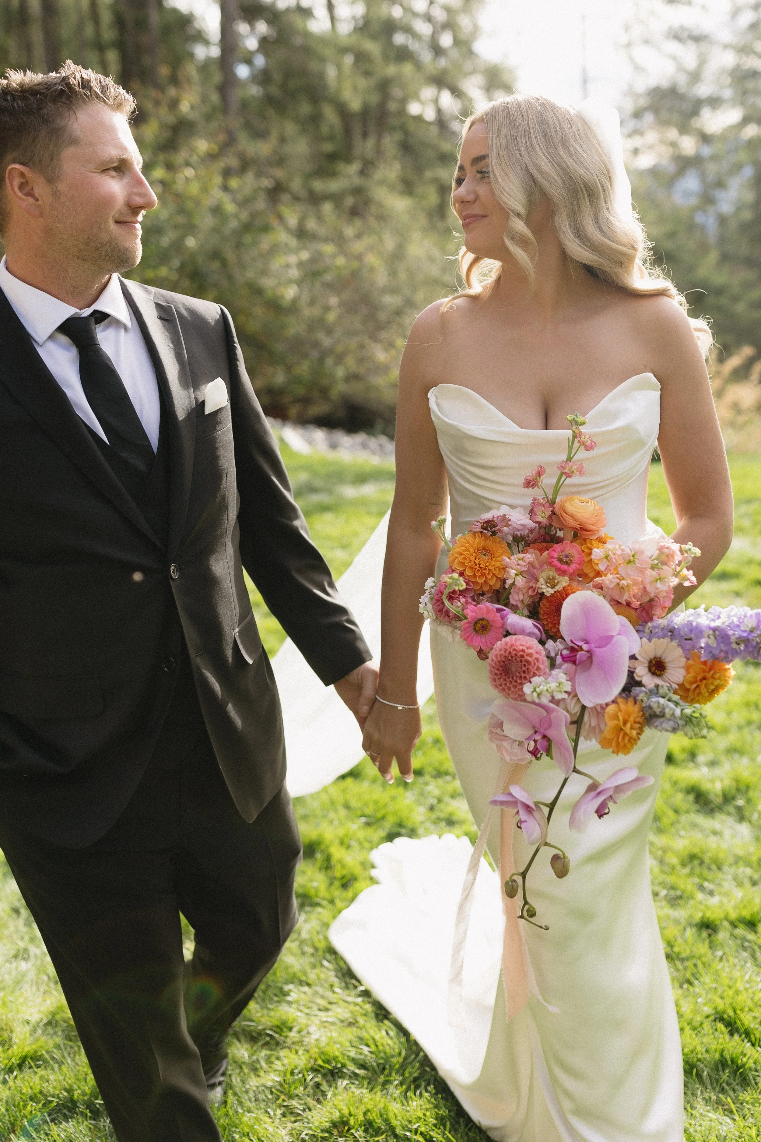 A bride and groom holding hands in a park, with the bride holding a large bouquet of pink, orange, and purple flowers, and the groom wearing a black suit and tie.