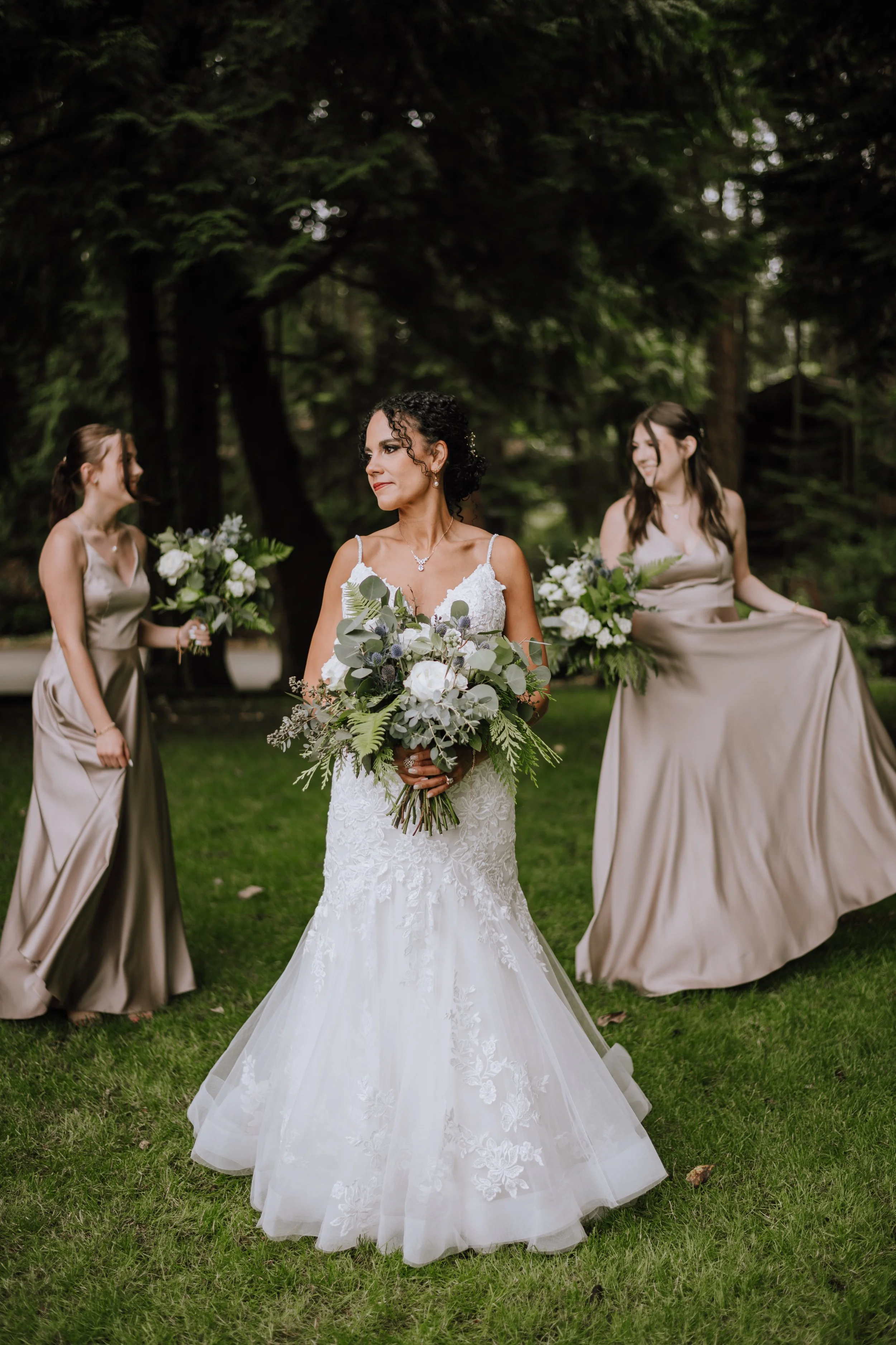 A bride in a white lace wedding dress holding a large bouquet, standing outdoors with two bridesmaids with bouquets in hand, in a green wooded setting.