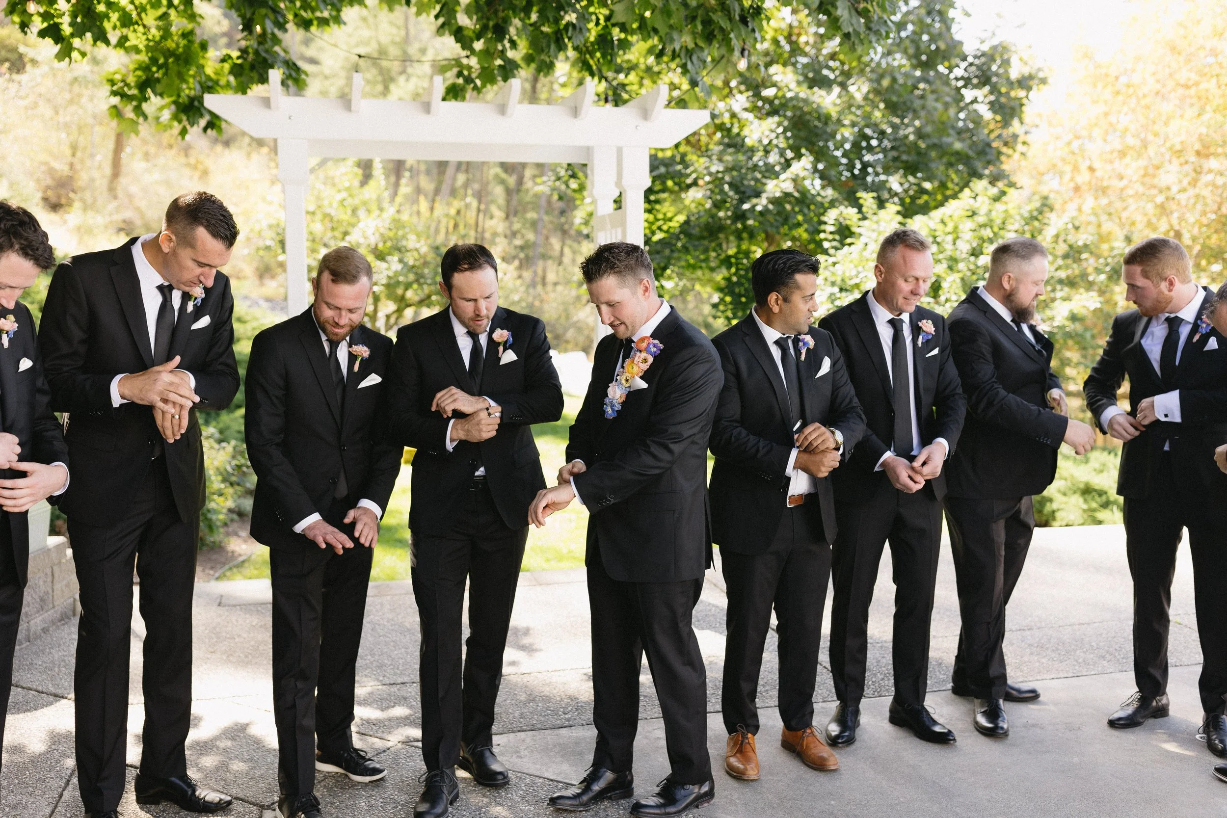 Groom and groomsmen dressed in black tuxedos with pink and purple boutonnières, standing outdoors on a ceremony day, adjusting their cuffs and watches, under a white arbor with a lush green background.