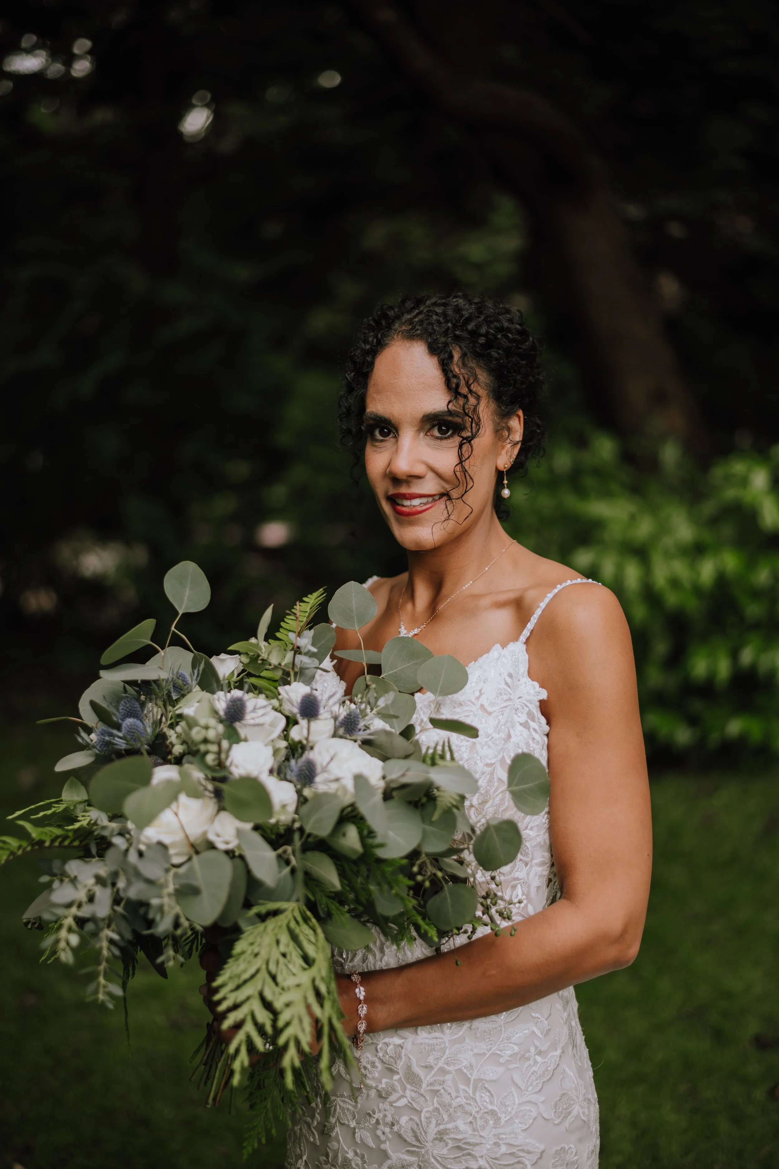 A woman in a white lace wedding dress holding a bouquet of white and purple flowers and green foliage standing outdoors in a lush garden.