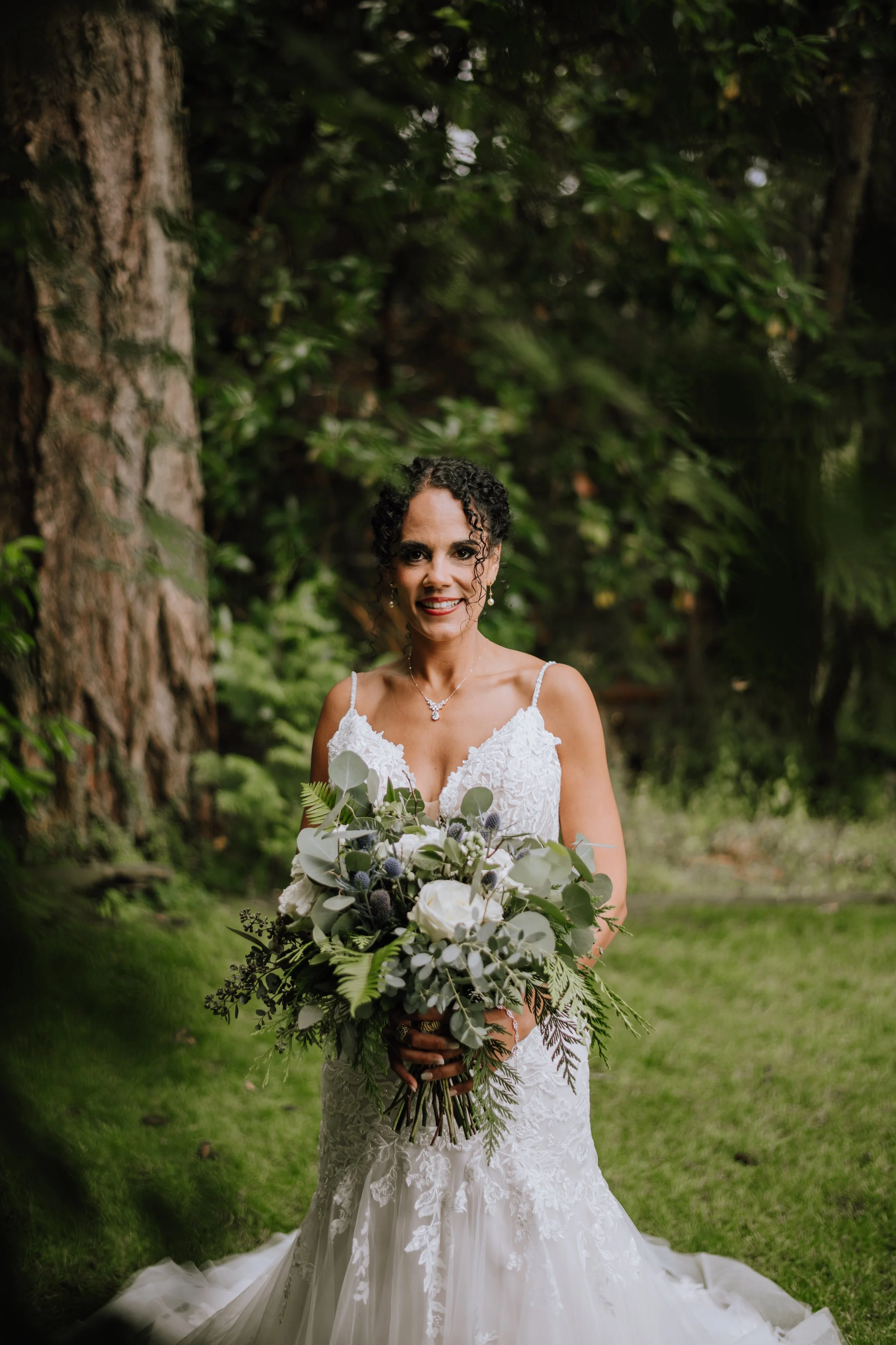 A bride in a white lace wedding dress holding a bouquet of white roses, greenery, and thistle, standing outdoors among trees and grass, smiling at the camera.