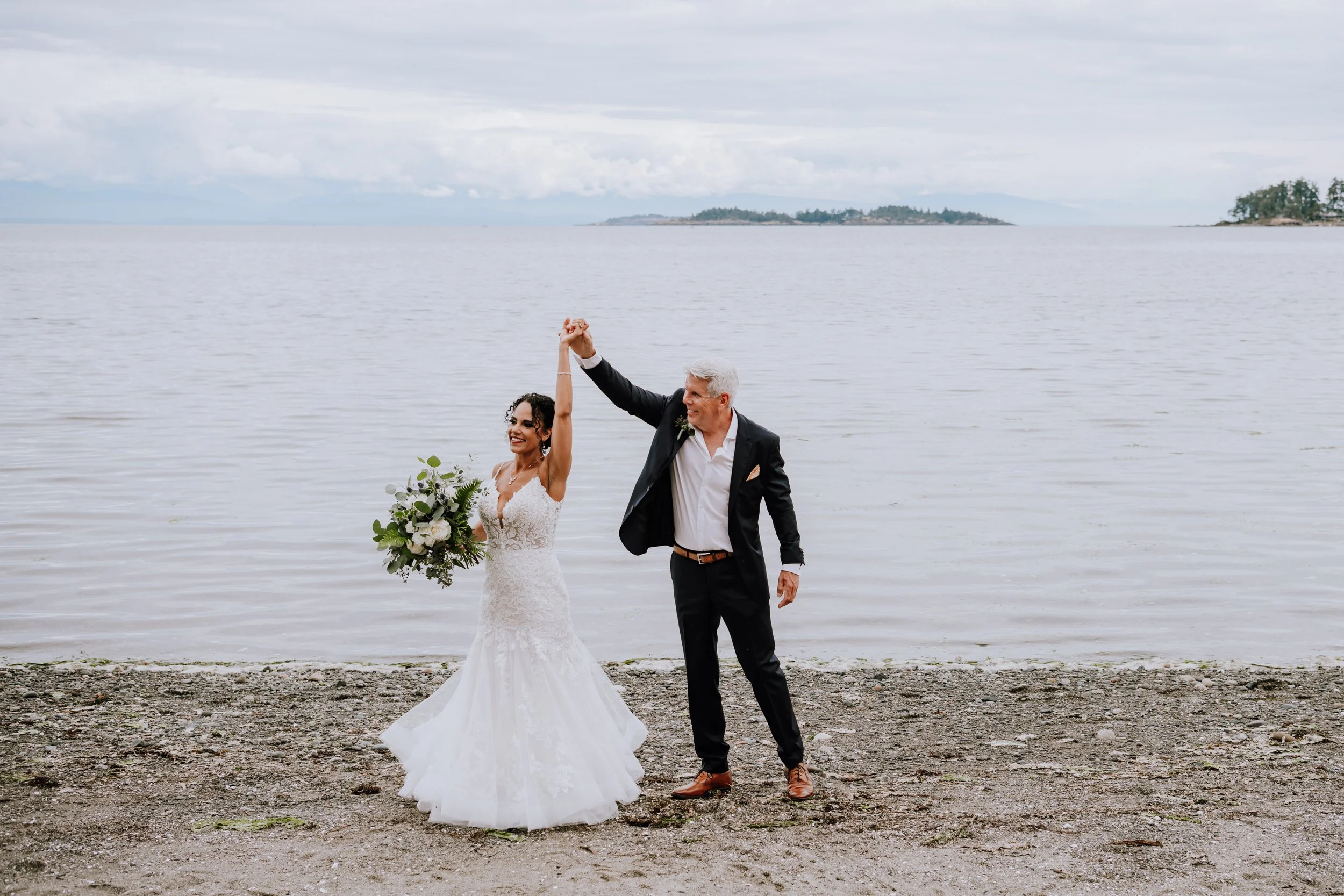 Bride and groom celebrating on a beach, with the bride holding a bouquet of flowers and smiling, as the groom raises her hand in victory. The scene includes water and distant islands in the background.