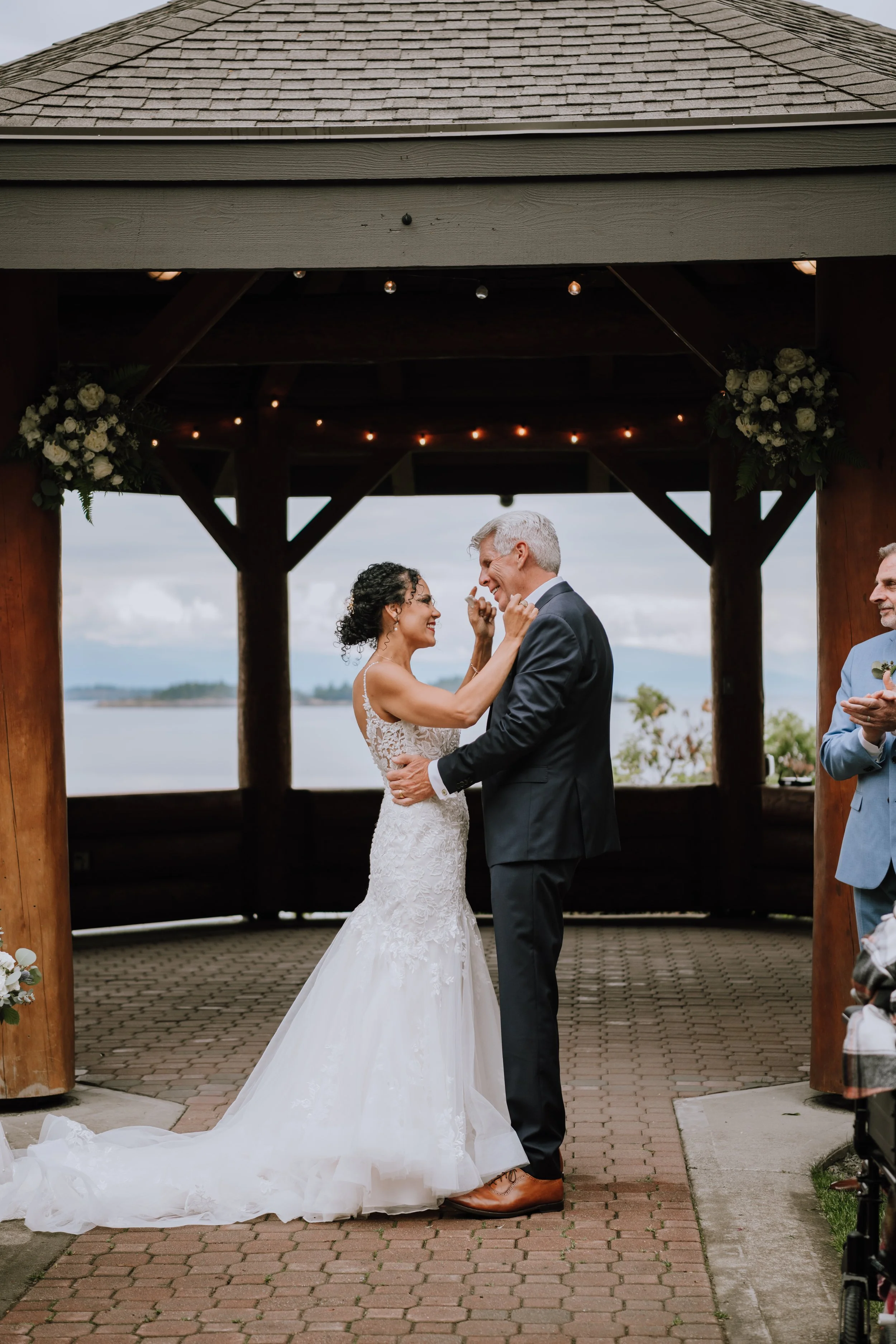Bride and groom sharing a dance at their wedding ceremony under a wooden gazebo overlooking the water, with wedding decorations and an officiant clapping nearby.